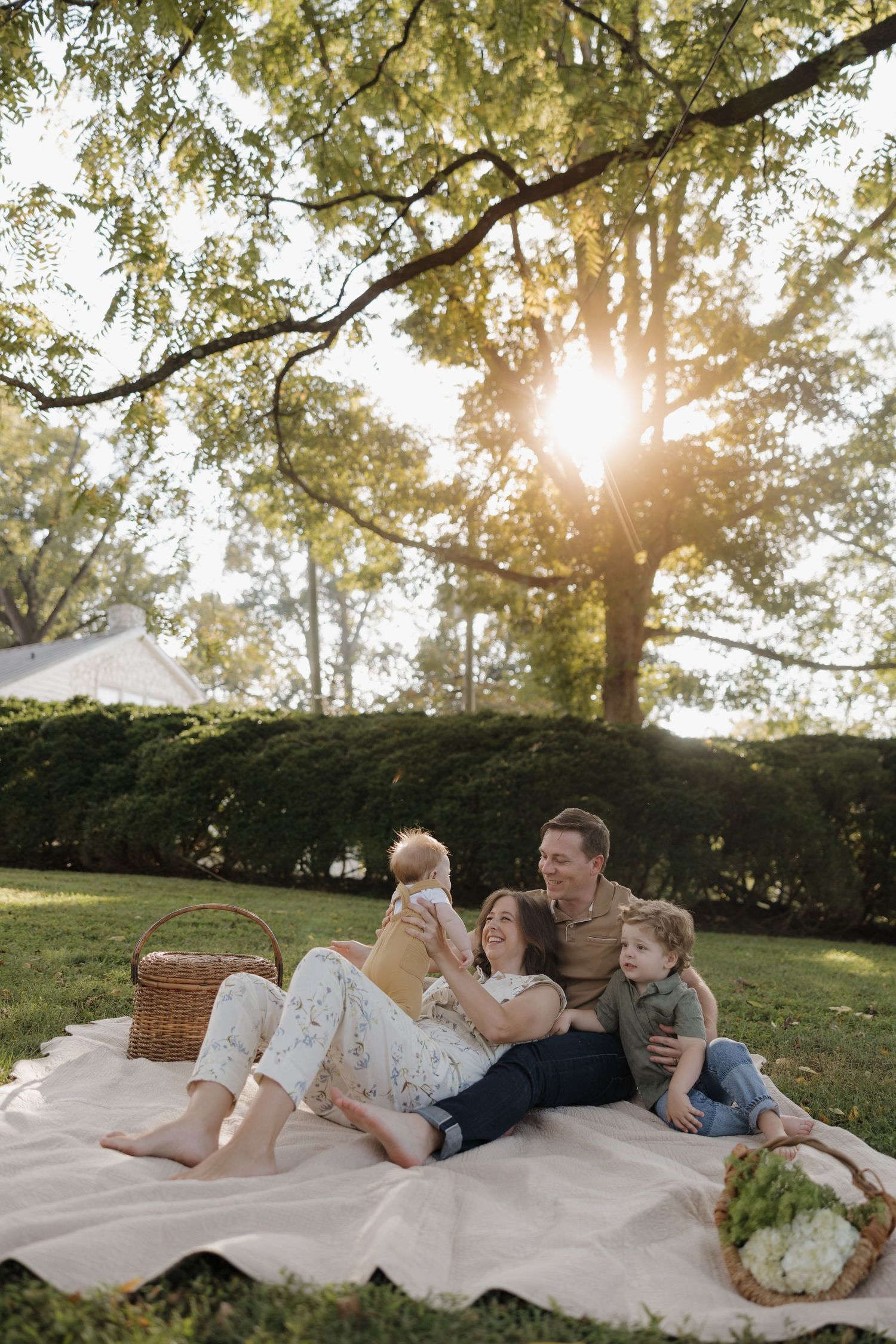 Family enjoying a picnic on a white blanket under sunlit trees in a park setting.