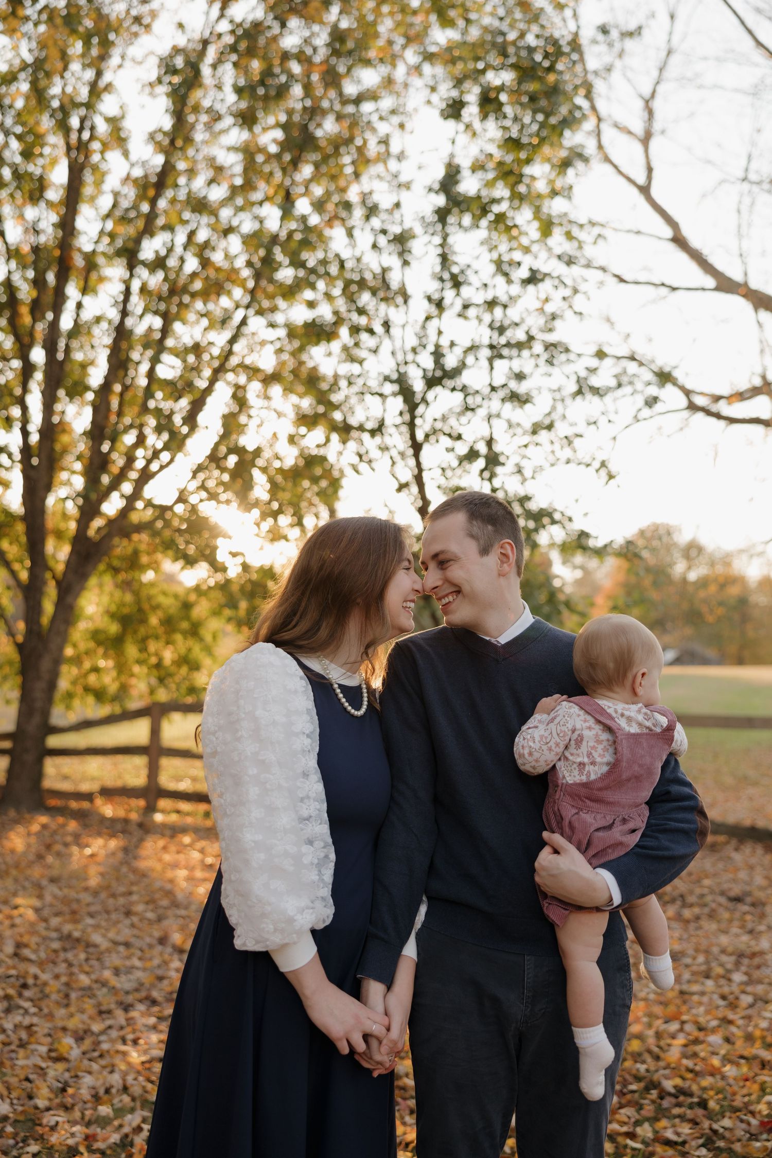 A couple shares a tender moment holding hands while the man cradles their baby girl in an autumn park setting.