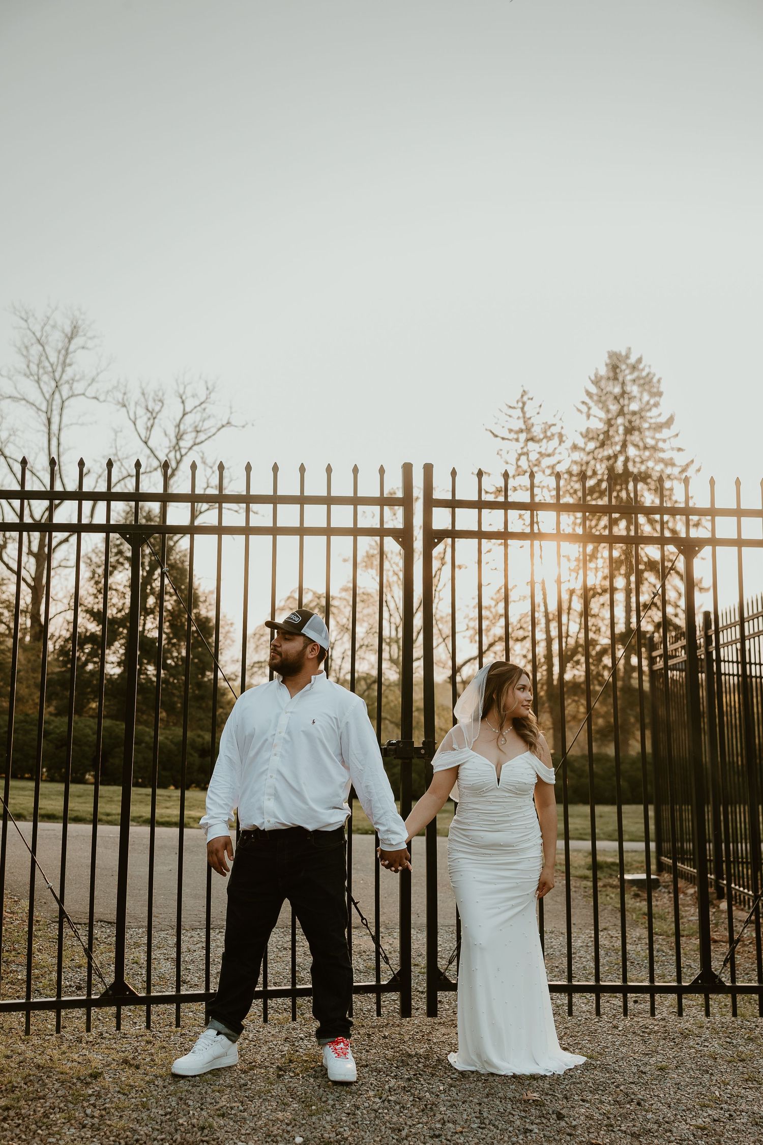 Couple in wedding attire poses against iron fence during sunset.