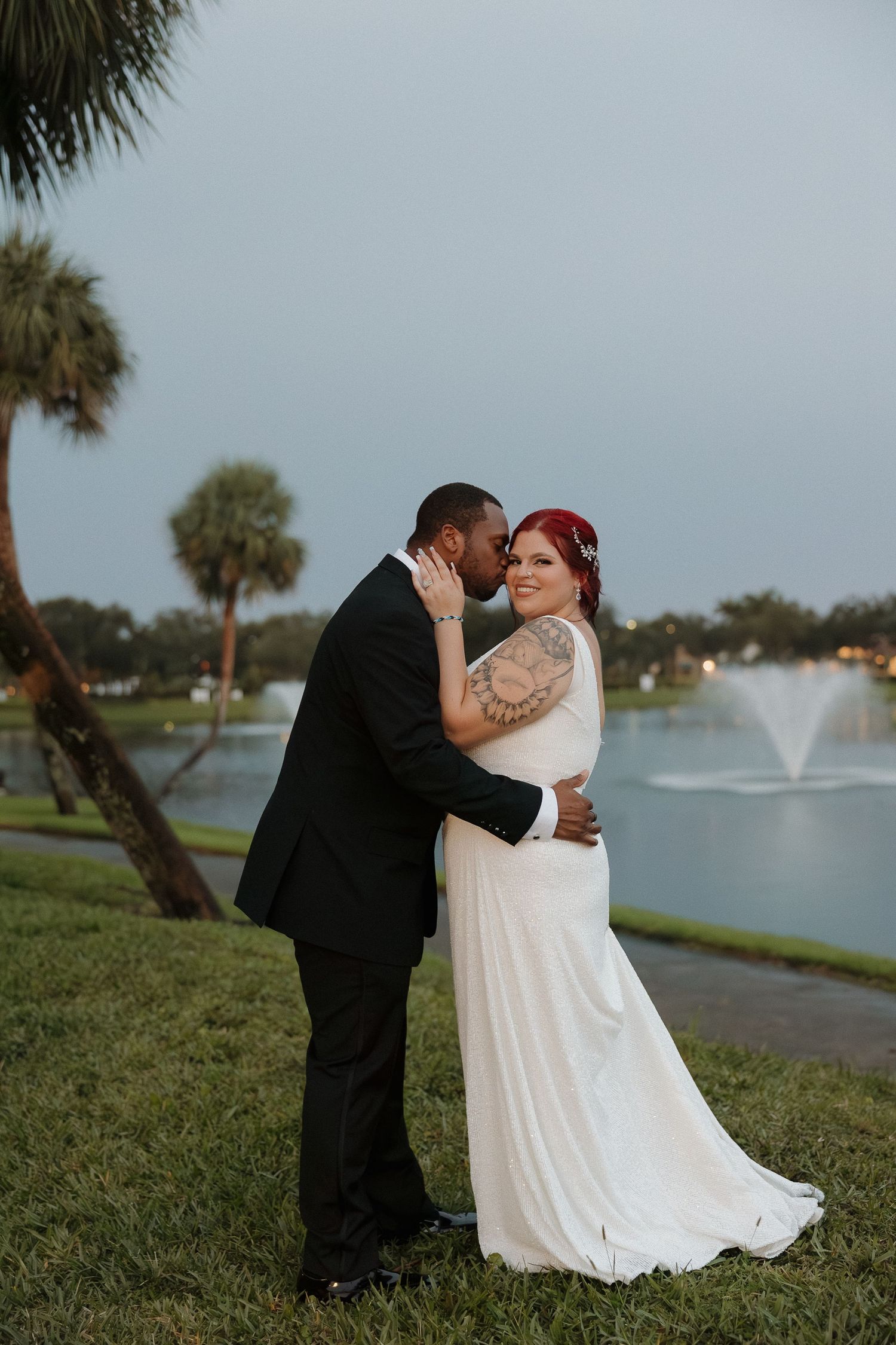 Wedding couple embracing near fountain with palm trees at sunset.