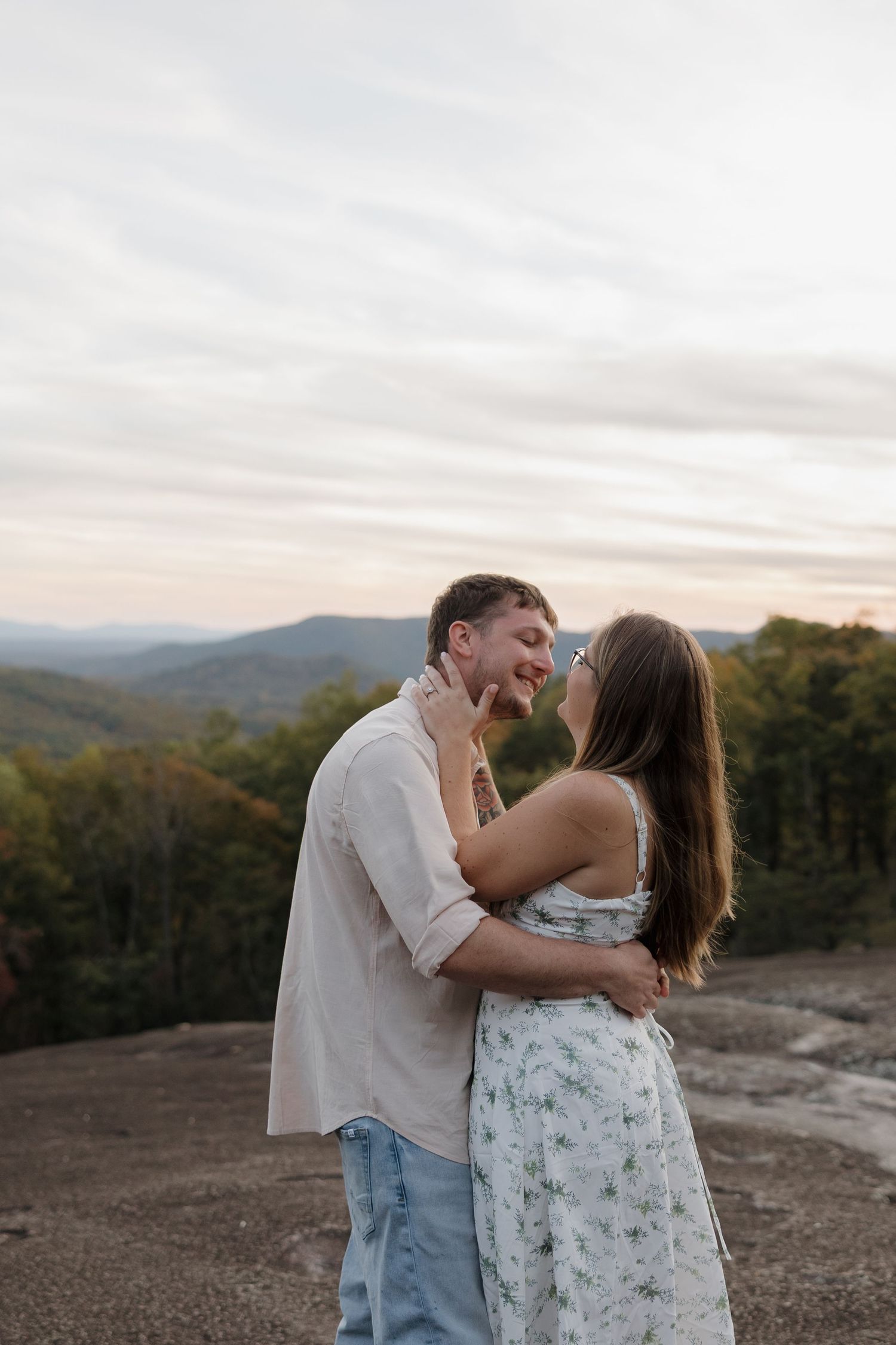 A romantic couple shares a joyful moment together on a mountaintop at sunset with rolling hills in the background.
