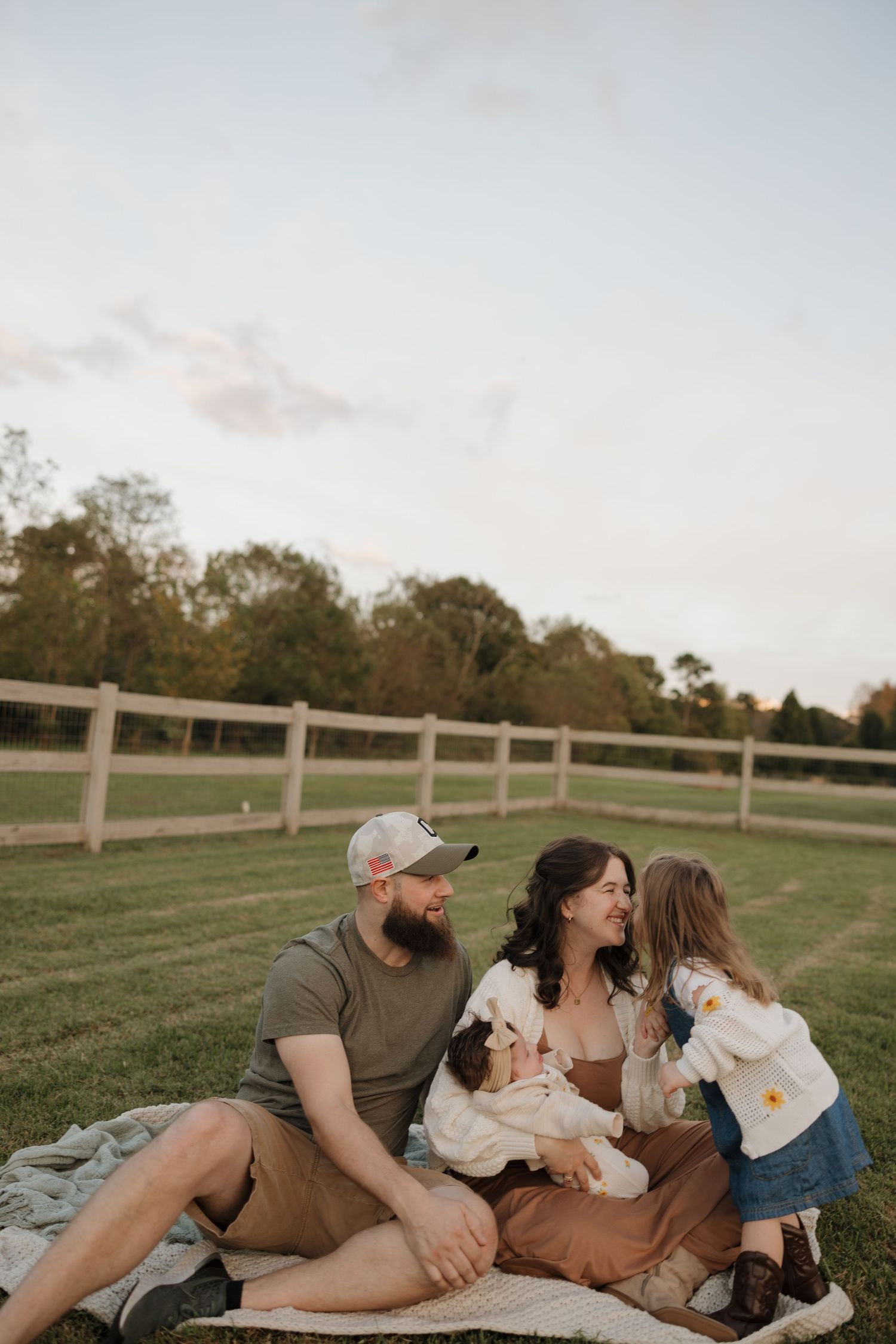 A family sits on a blanket with their small white dog on a grassy lawn with white fencing in the background.