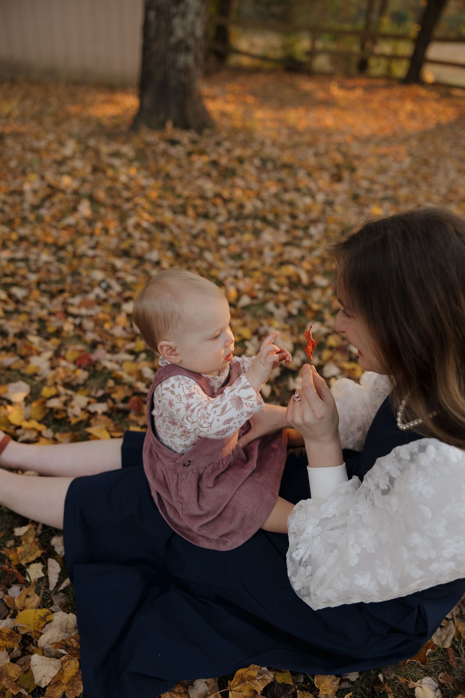 A parent and baby play together while lying on autumn leaves in a park setting.