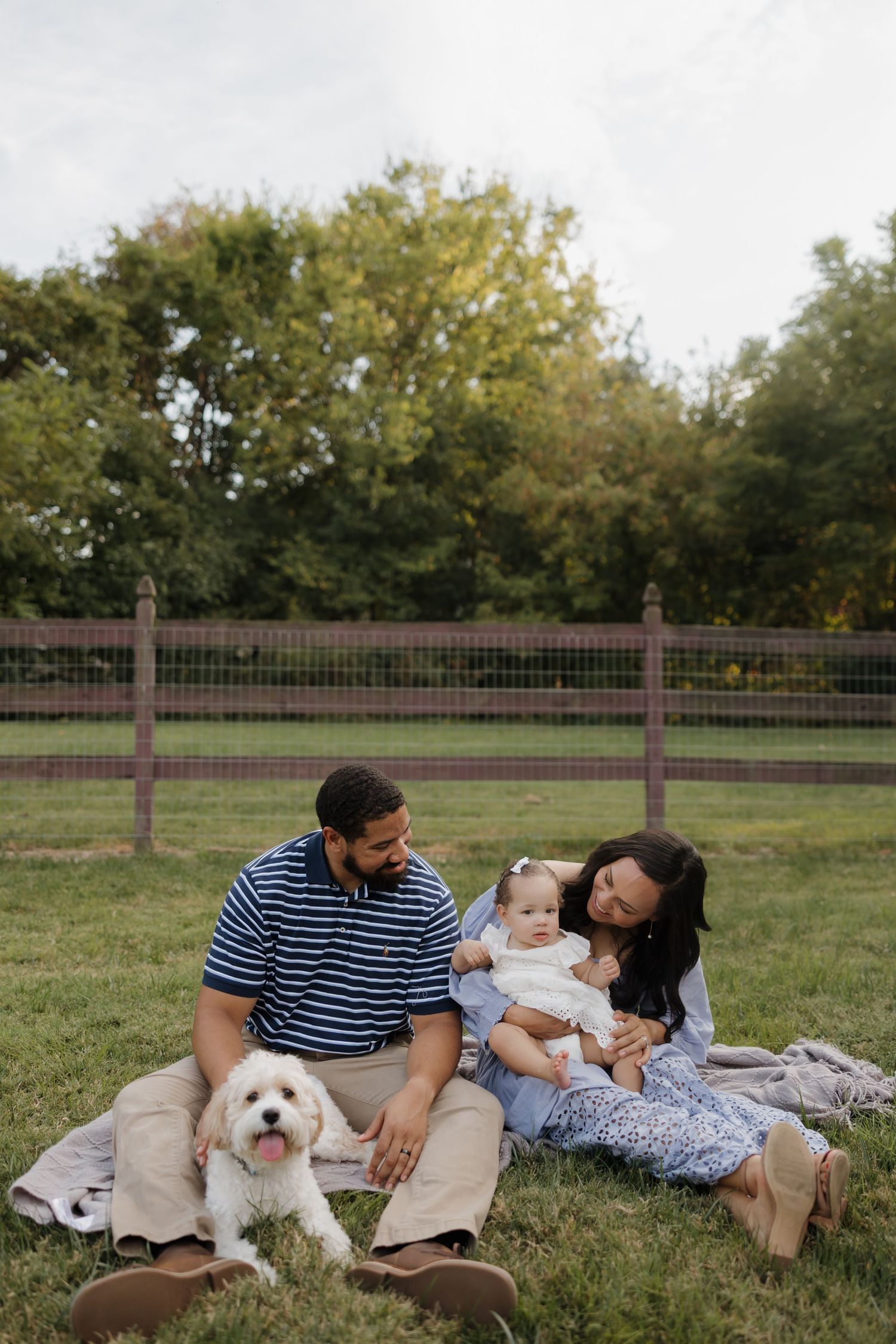 Family enjoying a relaxed afternoon picnic on grass with their white fluffy dog near a wooden fence.