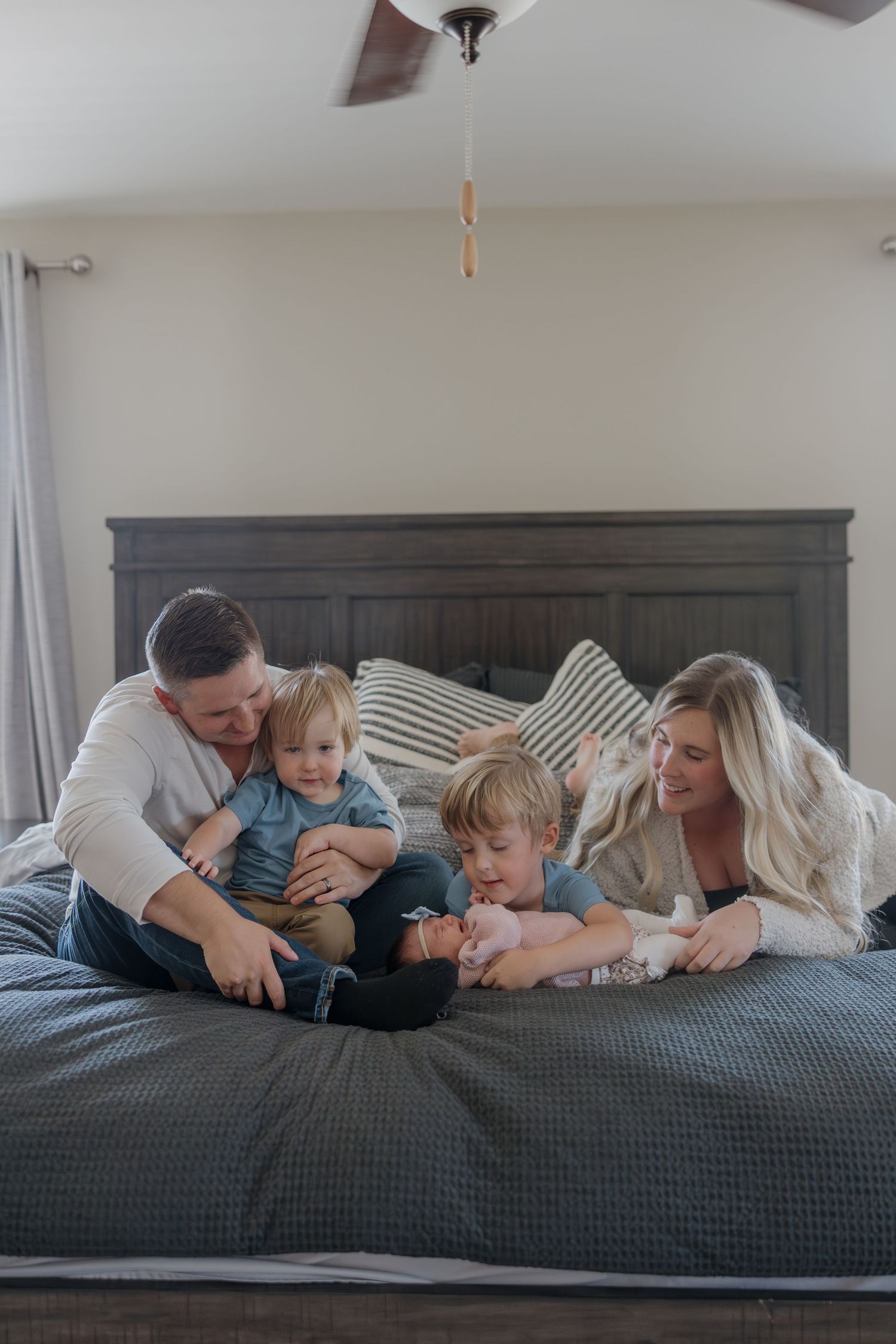 Family of four lying together on a gray bed sharing tender moments in a series of candid lifestyle photos.