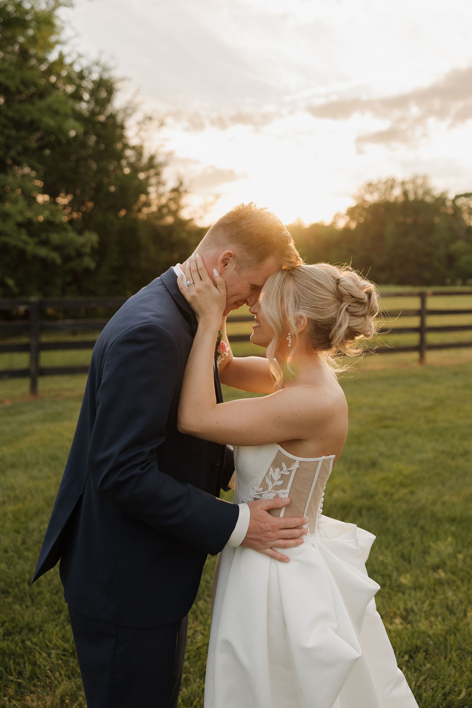 Bride and groom share a romantic sunset moment outdoors, with golden light glowing behind them near a wooden fence.
