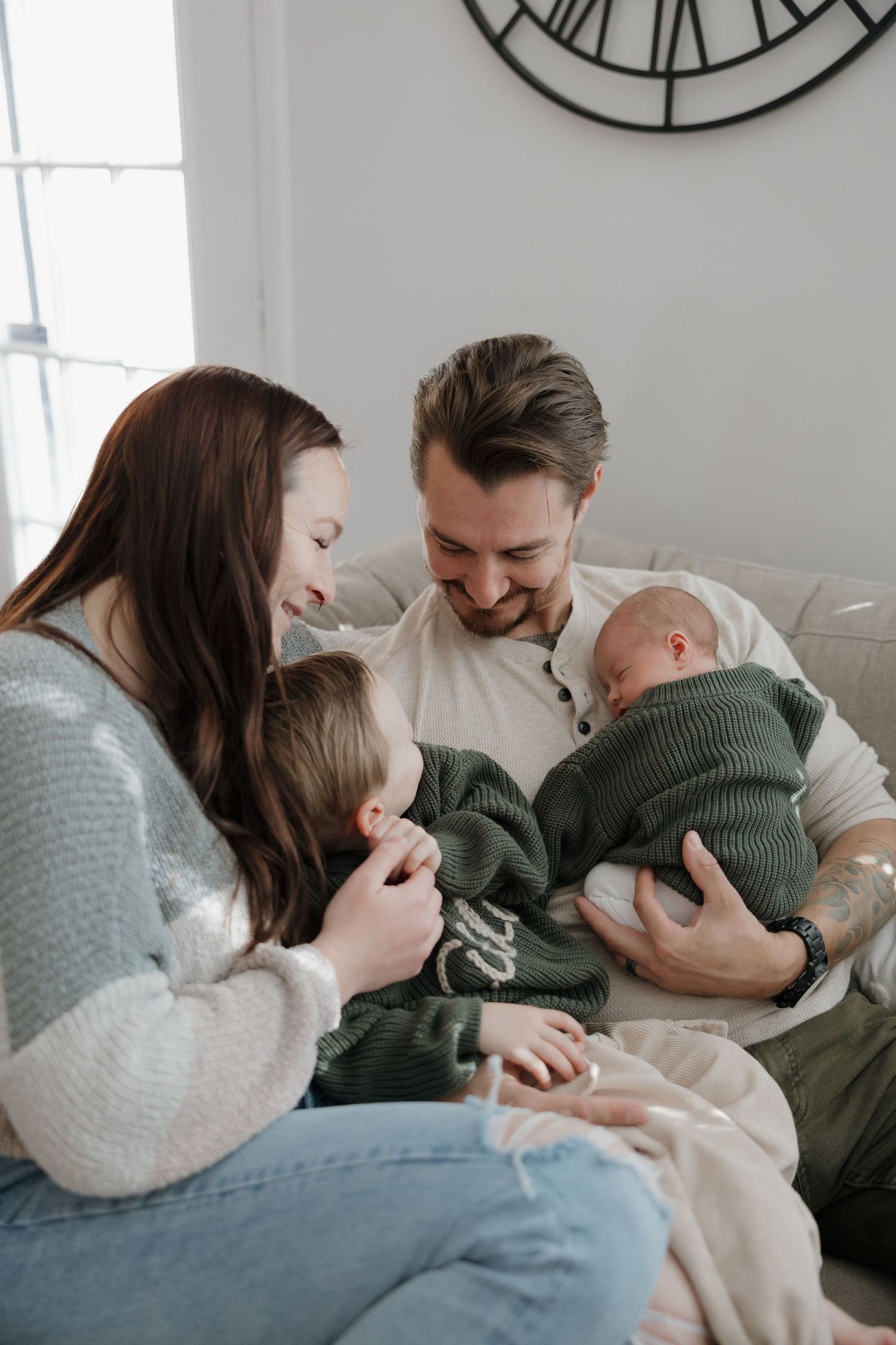 A happy family of four sits together on a couch, parents smiling at their newborn baby while a toddler looks on.