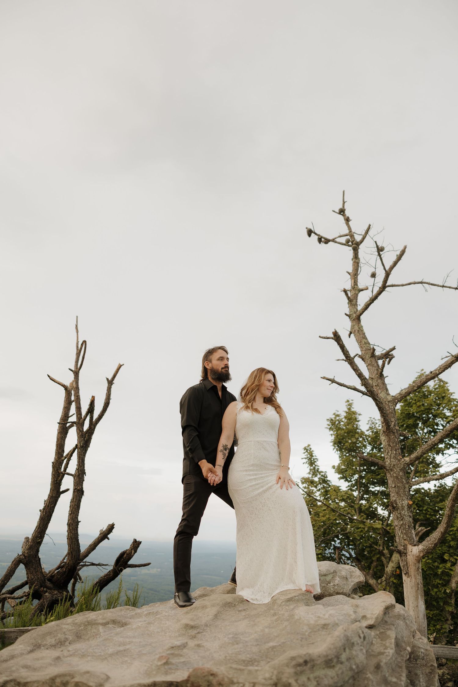 A couple stands together on a rocky mountain ledge with bare trees framing the scenic view.
