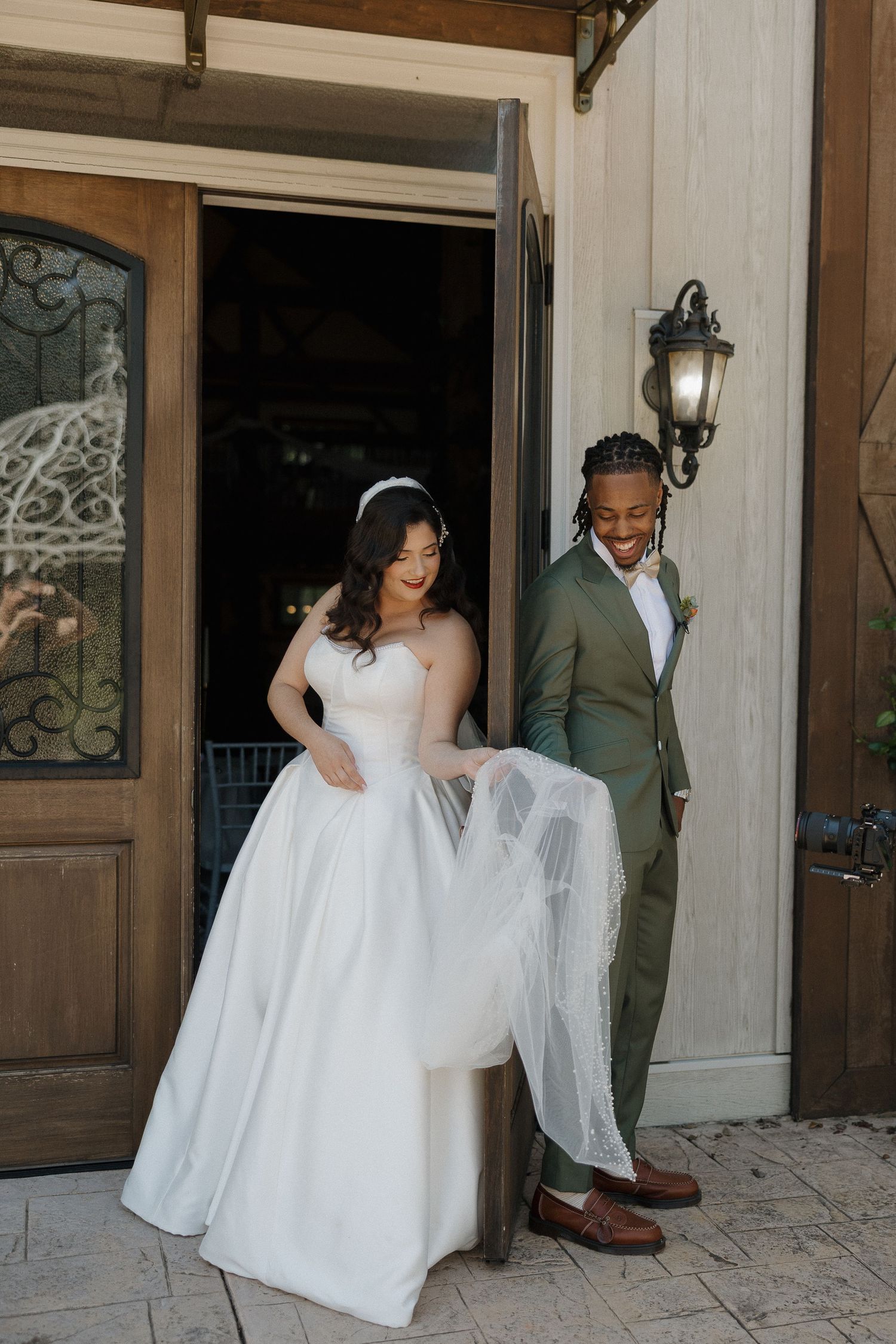 Joyful couple in wedding attire standing by elegant wooden doorway.