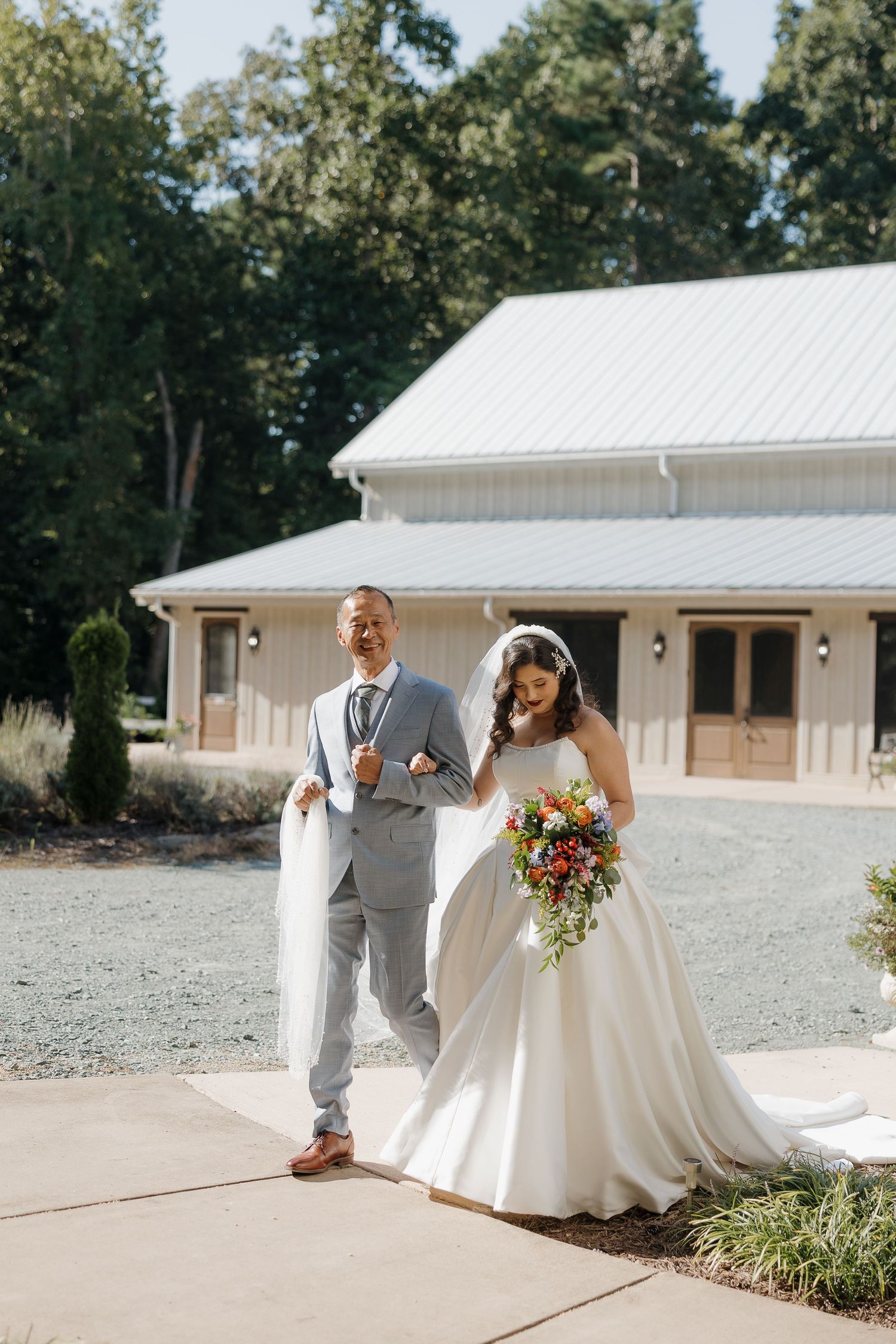 A joyful wedding couple walks outside a modern white barn venue with the bride holding a vibrant bouquet.