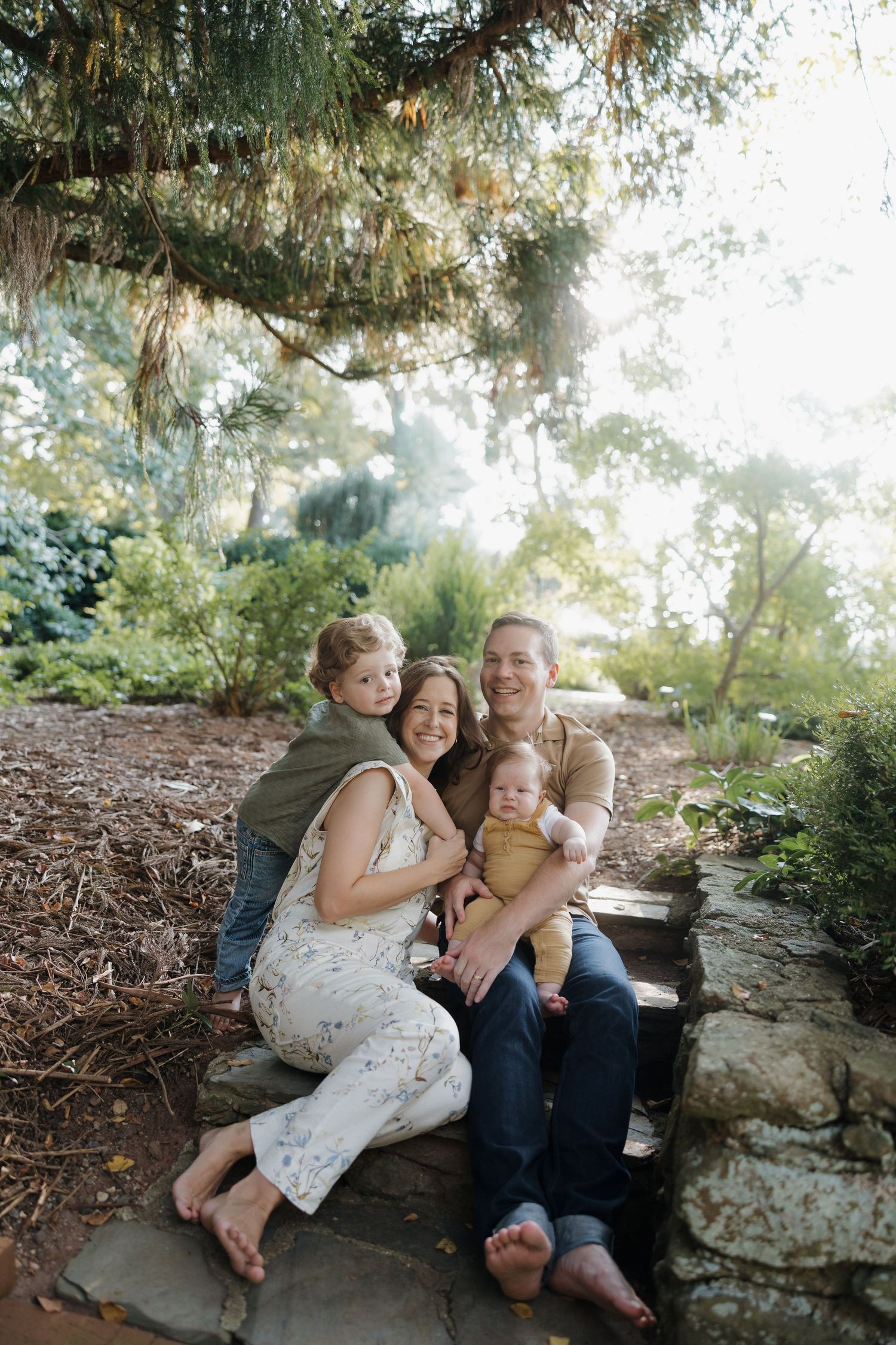A family sits together on stone steps surrounded by lush greenery and dappled sunlight in an outdoor setting.