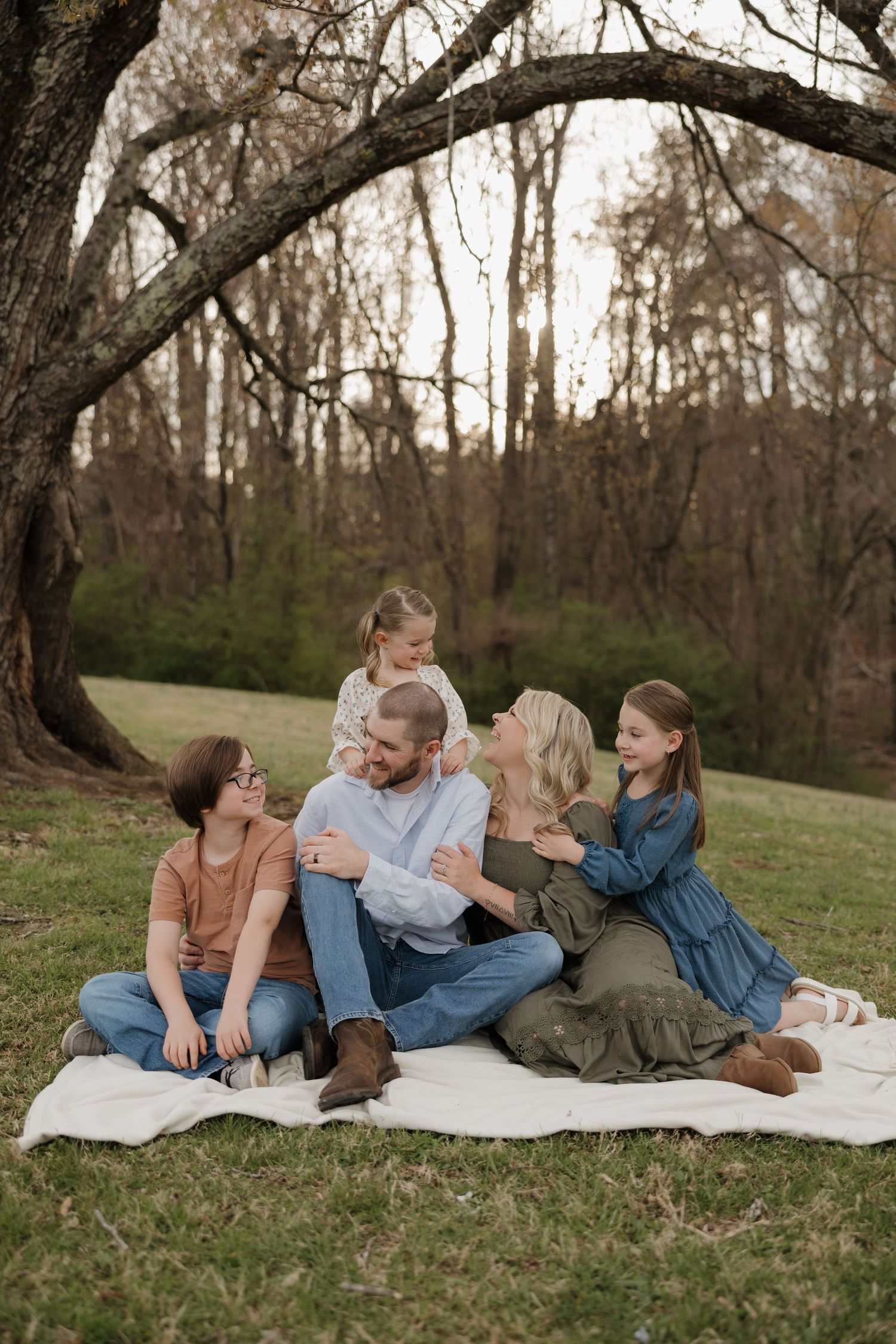 Family of five sitting on a blanket outdoors under a large tree, sharing a warm embrace in a lush green park.