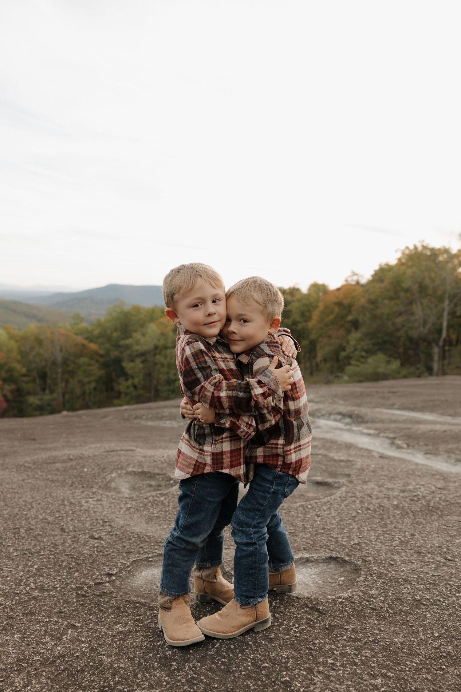 Two toddlers in matching plaid shirts and jeans share a sweet moment together on a rocky mountain overlook at sunset.