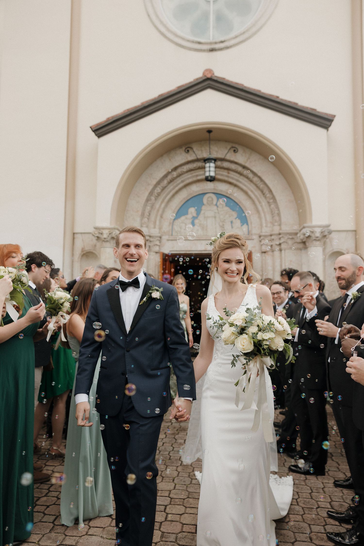 Wedding guests celebrate with bubbles outside a church with an arched entrance.