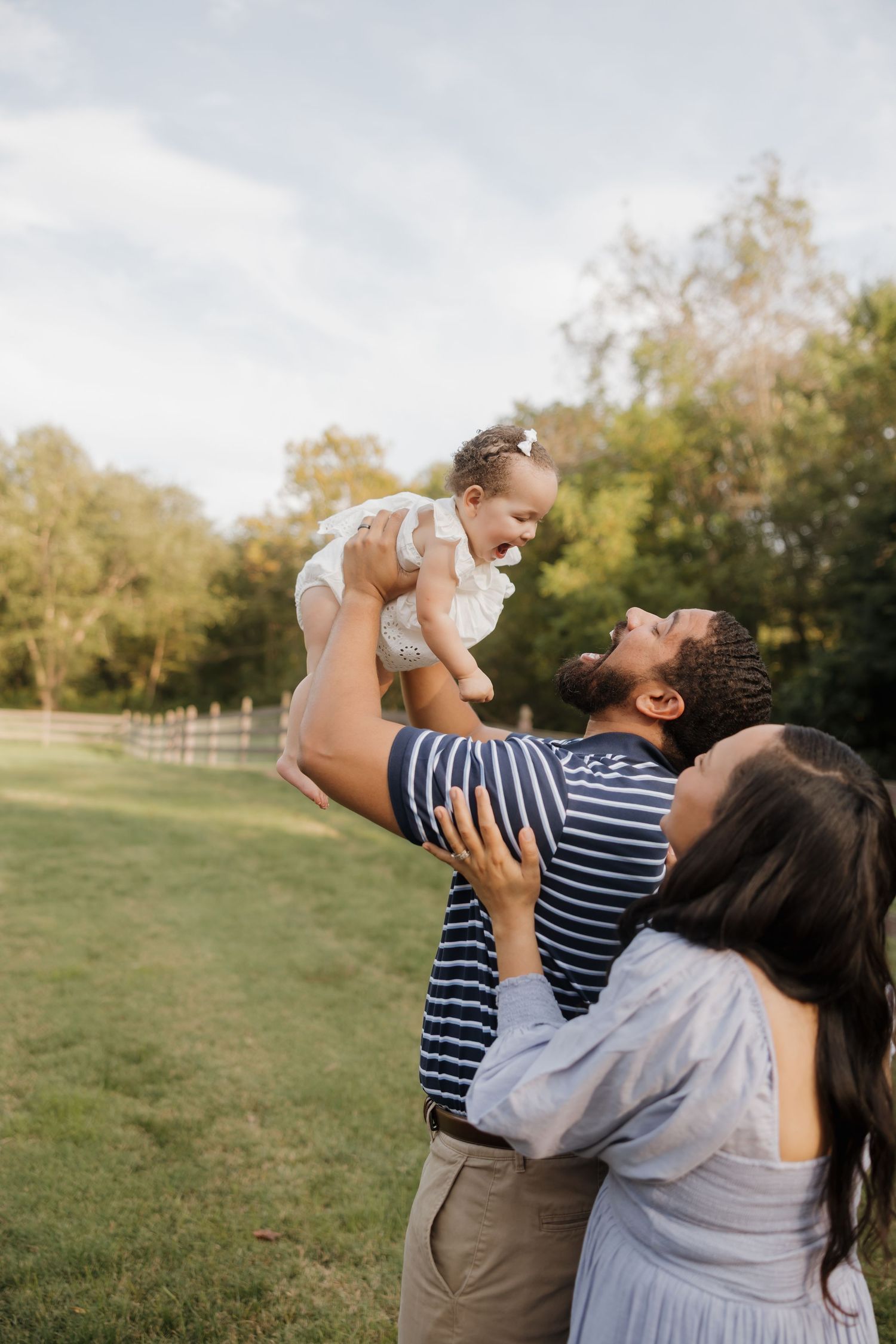 Happy family outdoors, father lifting baby girl in air while mother watches, green park setting.
