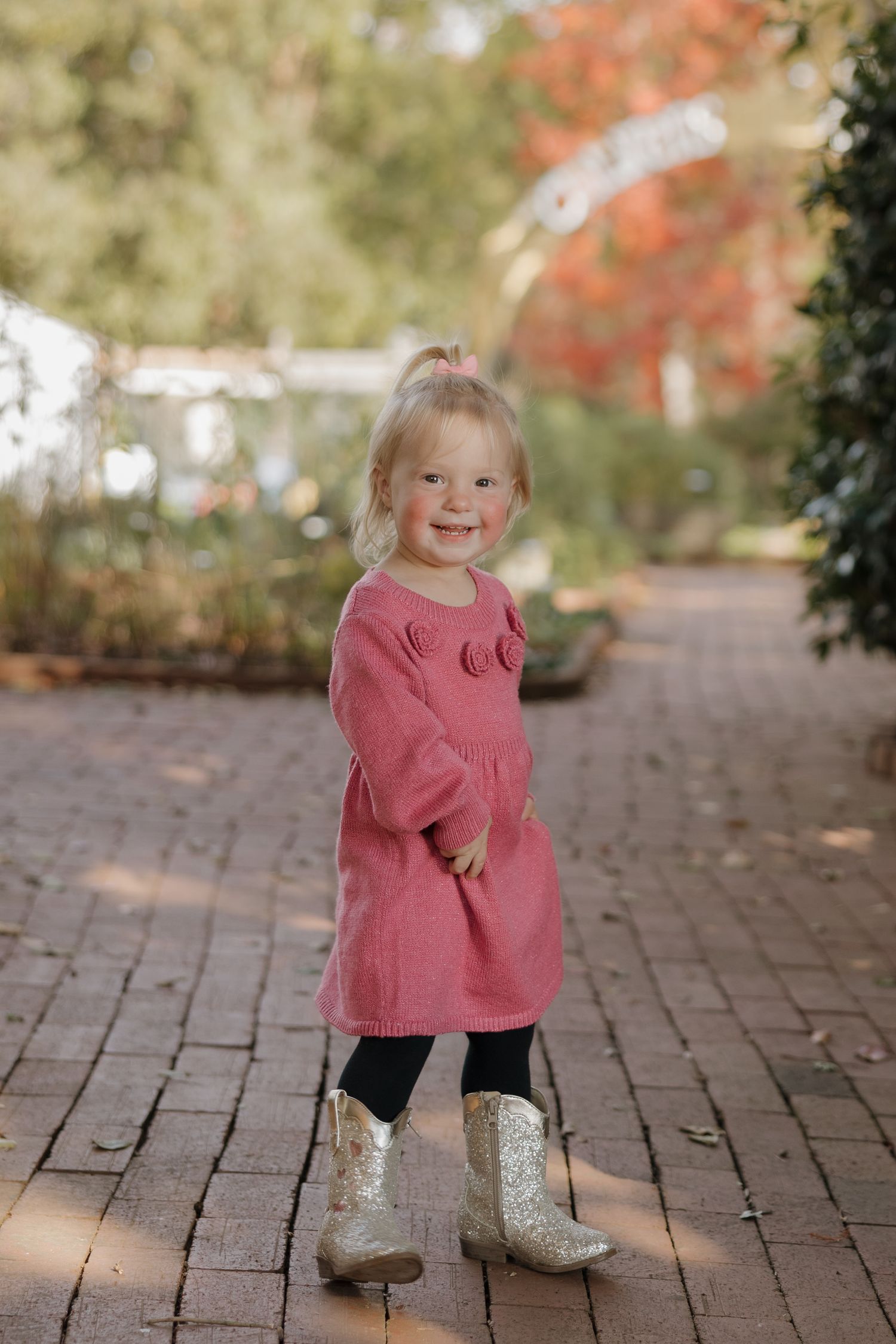 A child in a pink sweater and sparkly boots plays on a brick path surrounded by autumn foliage.