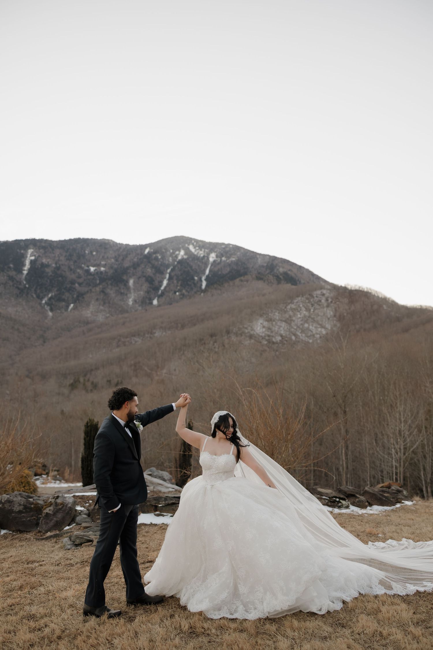 Bride in white gown and groom in black suit dance with mountain backdrop during outdoor winter wedding.