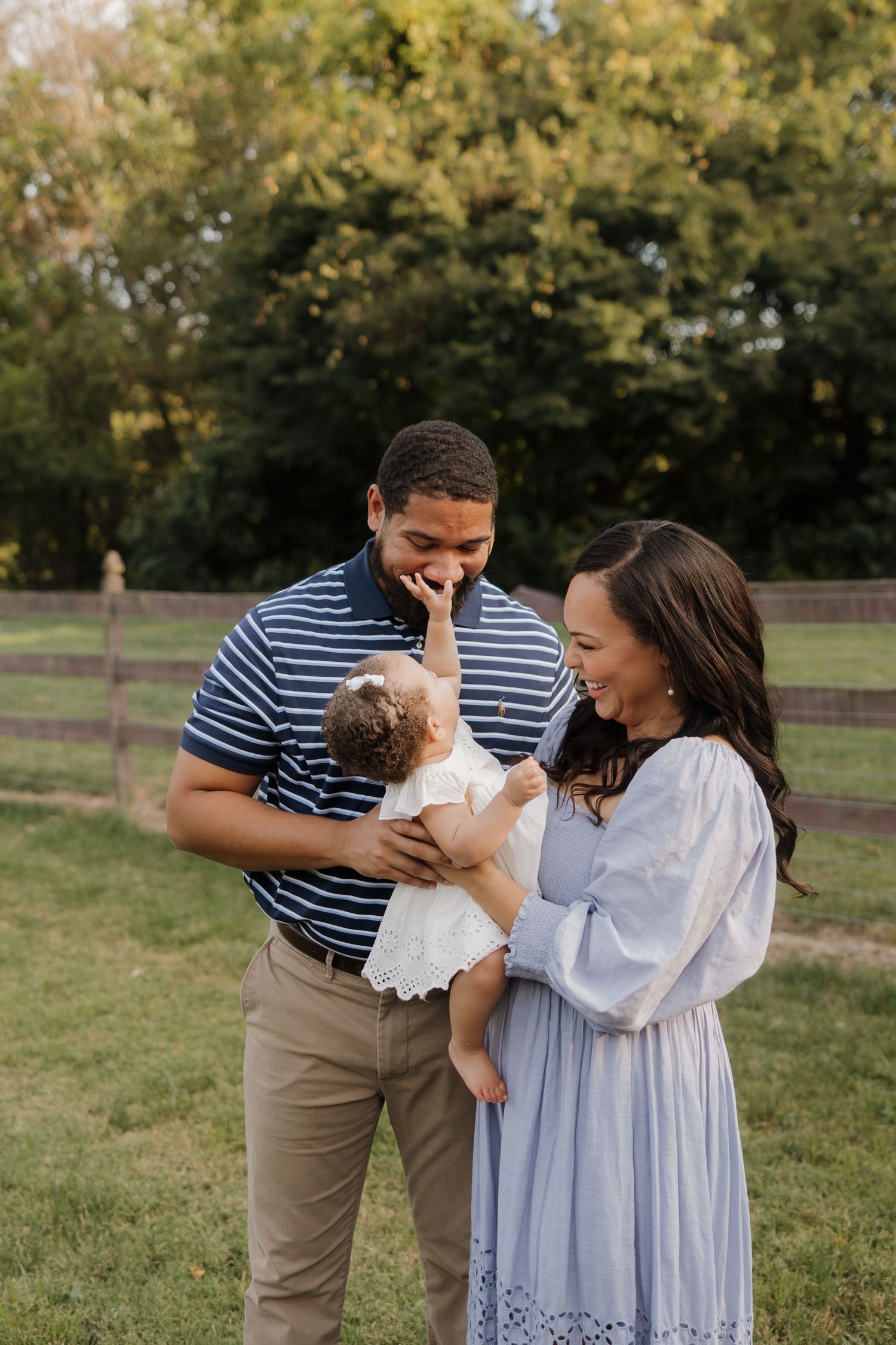 A couple in blue and white outfits share tender moments with their baby in a grassy outdoor setting at sunset.