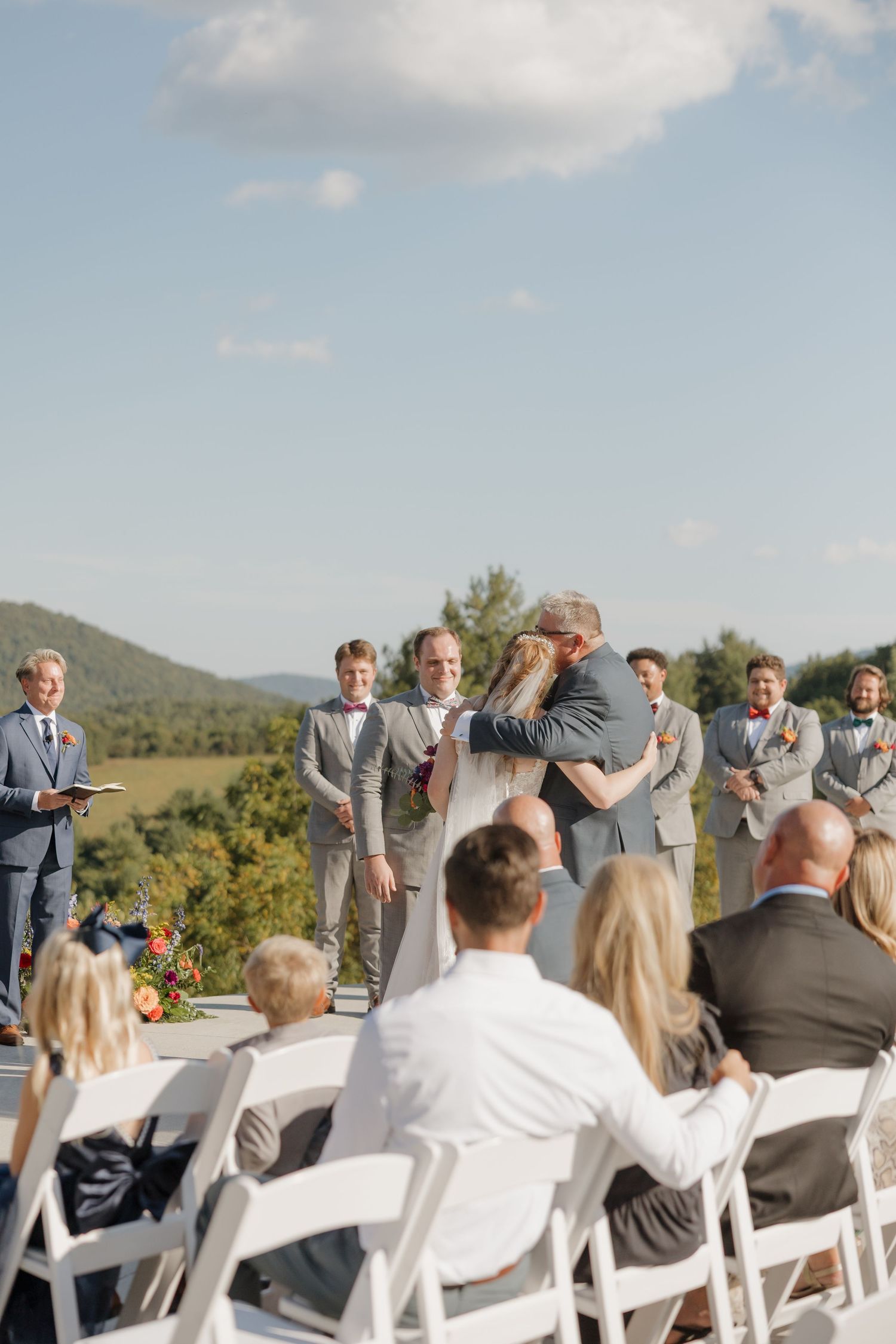 Outdoor wedding ceremony with guests seated in white chairs against mountain scenery.