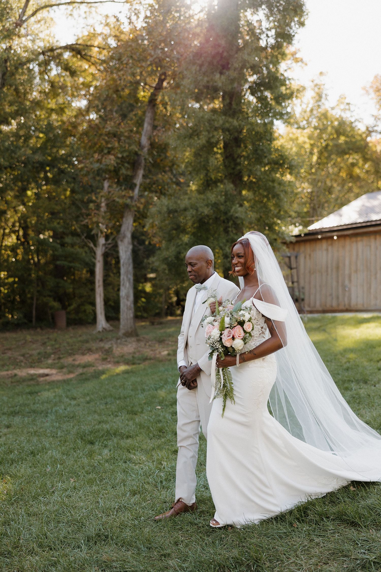 A couple in wedding attire poses outdoors on a grassy hill during golden hour.