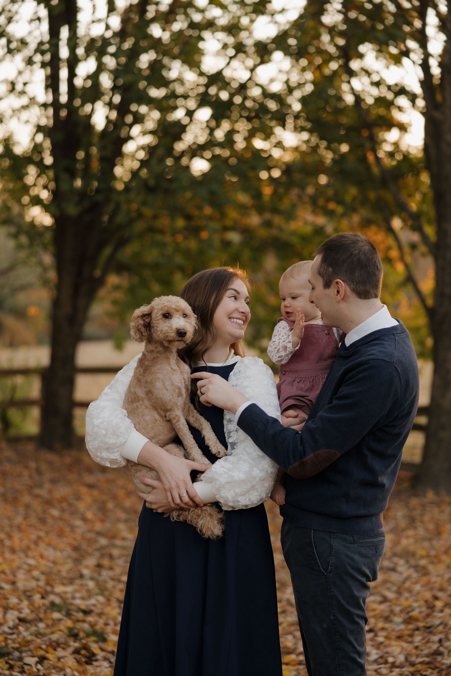 A couple plays with their small dog in a park covered with autumn leaves at sunset.