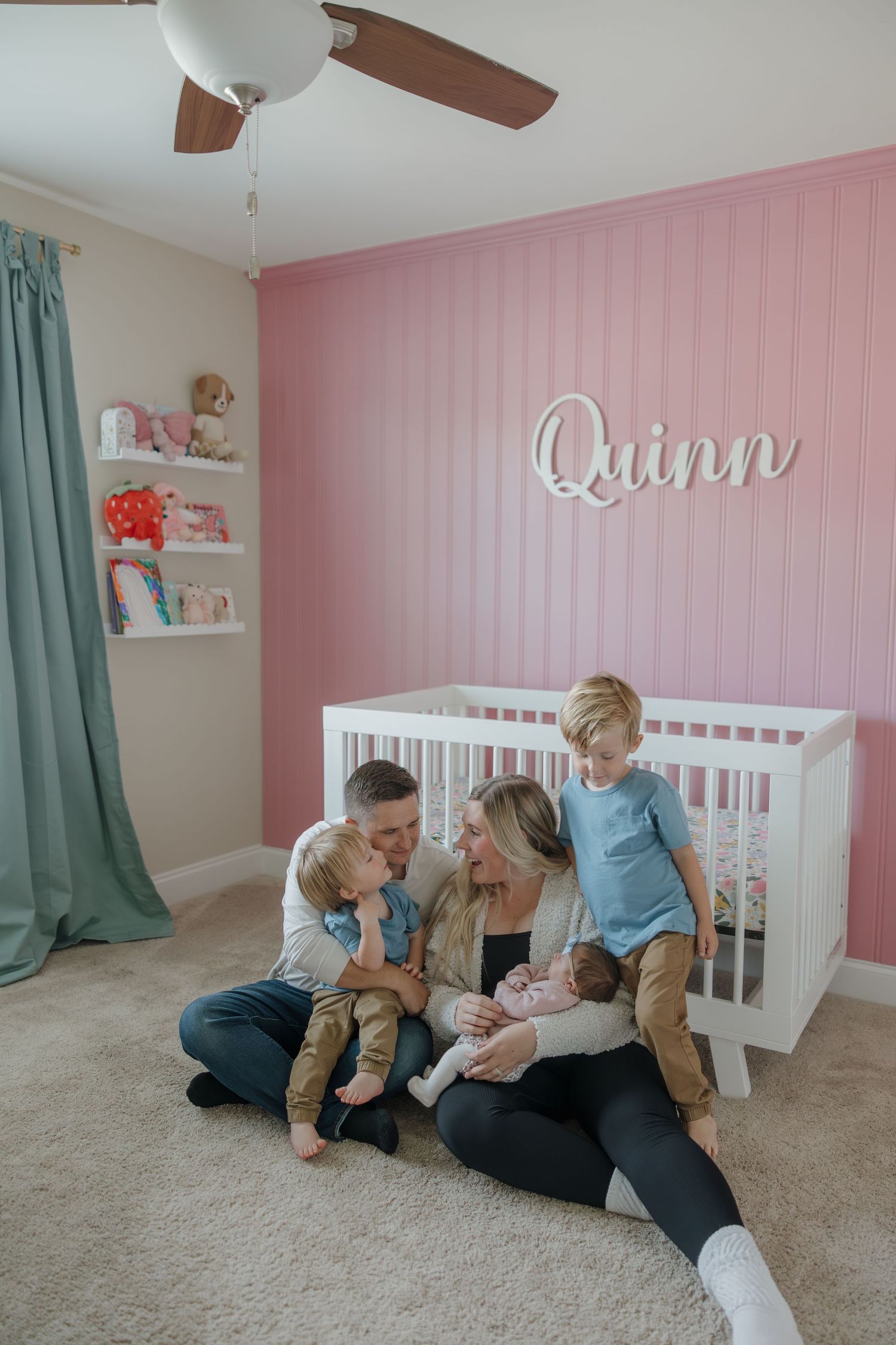 Nursery room with pink accent wall, white crib, teal curtains, floating shelves and name 'Quinn' mounted above the crib.