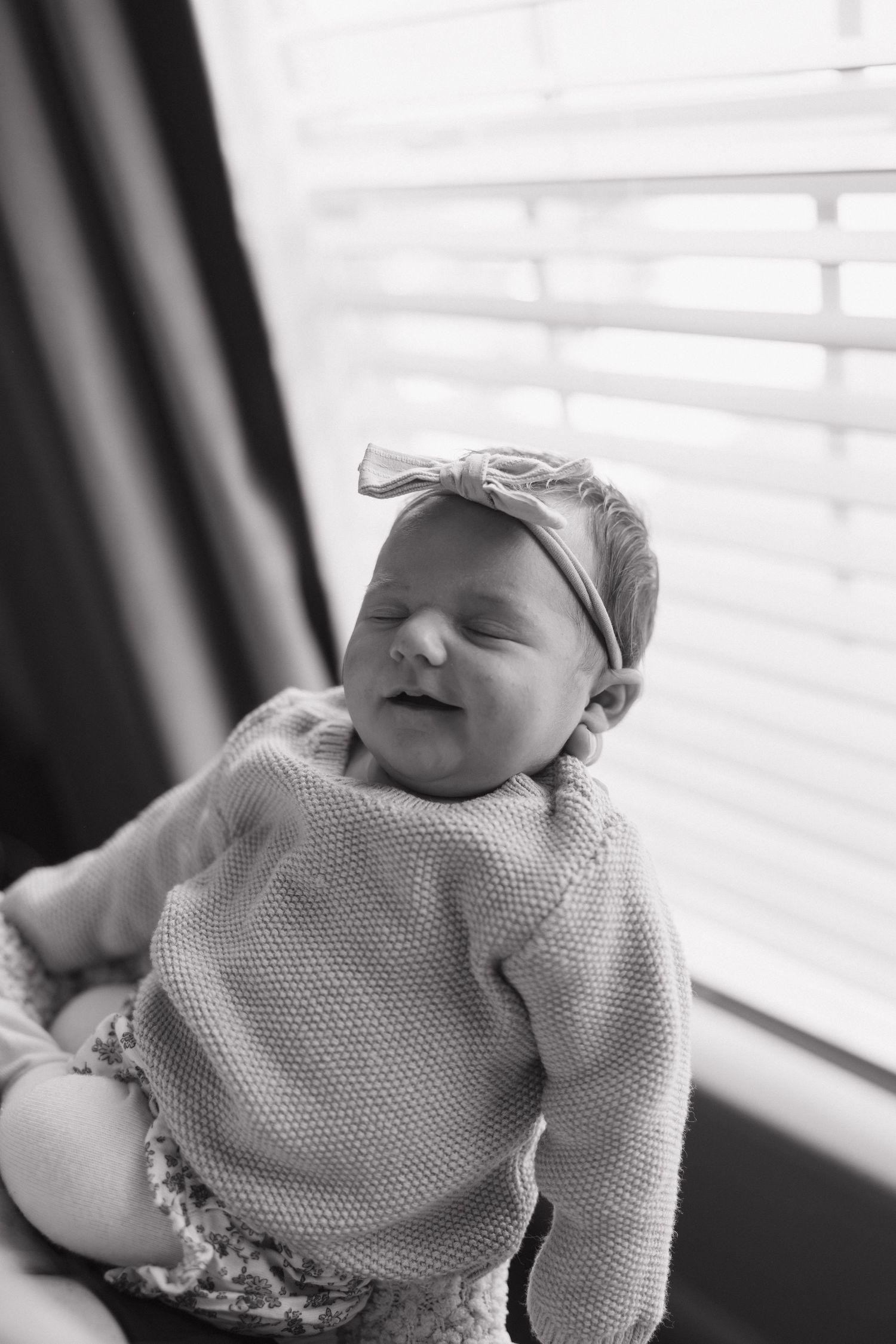Black and white photo of a sleeping newborn baby wearing a knit sweater against window light.