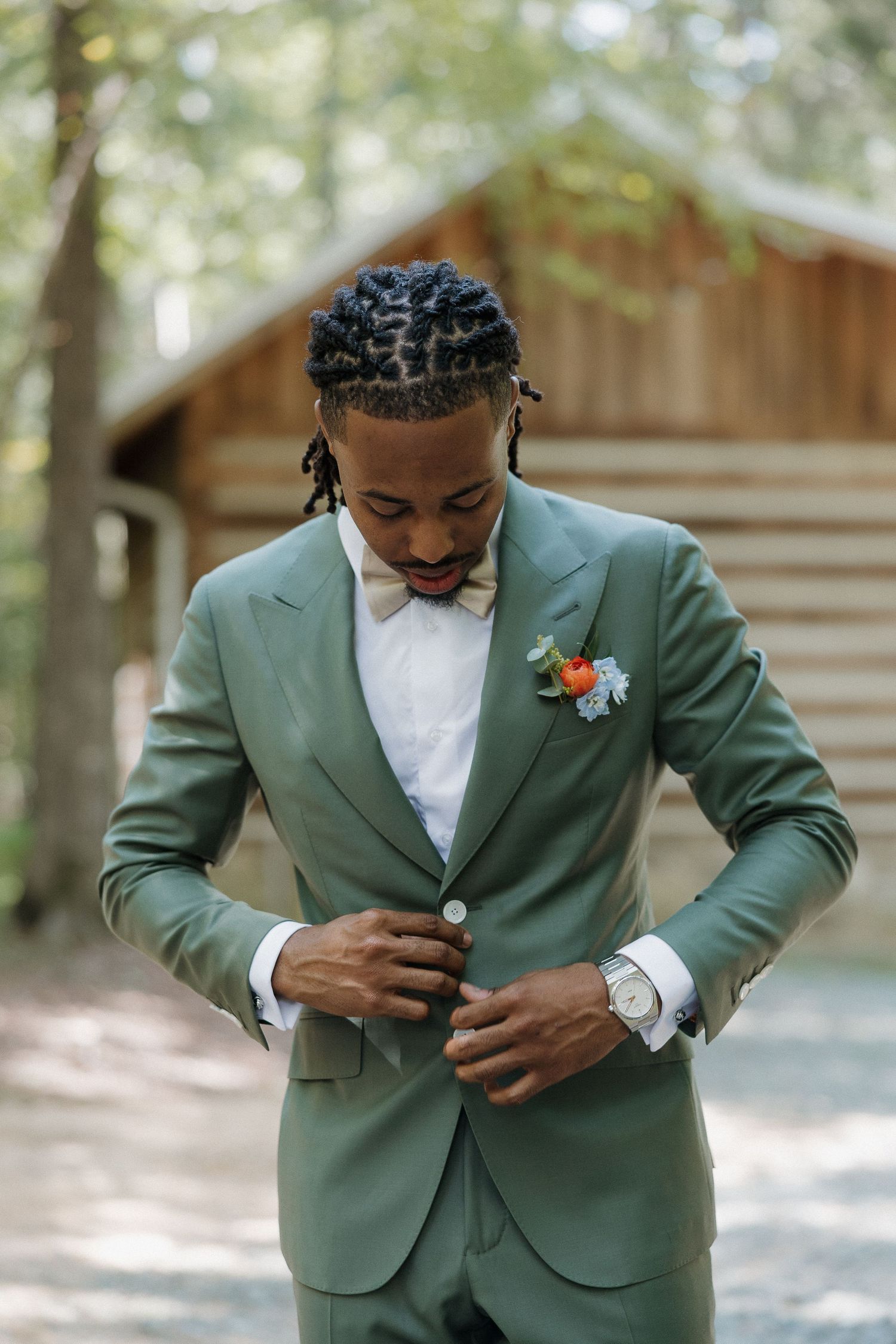 Groom in sage green suit adjusting jacket button in outdoor setting.