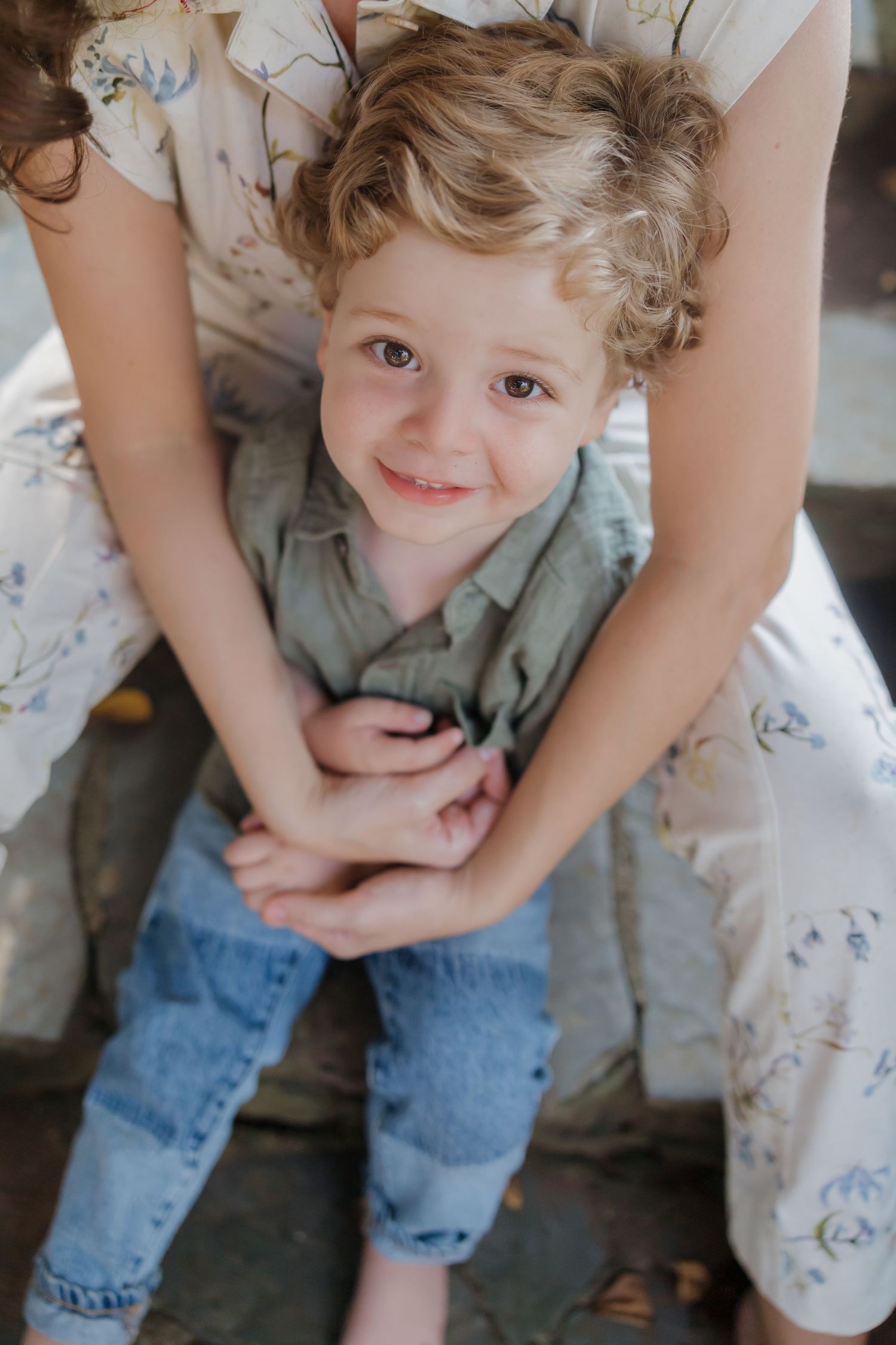 A young child with curly blonde hair wearing a mint green shirt and jeans sits in someone's lap wearing a floral dress.