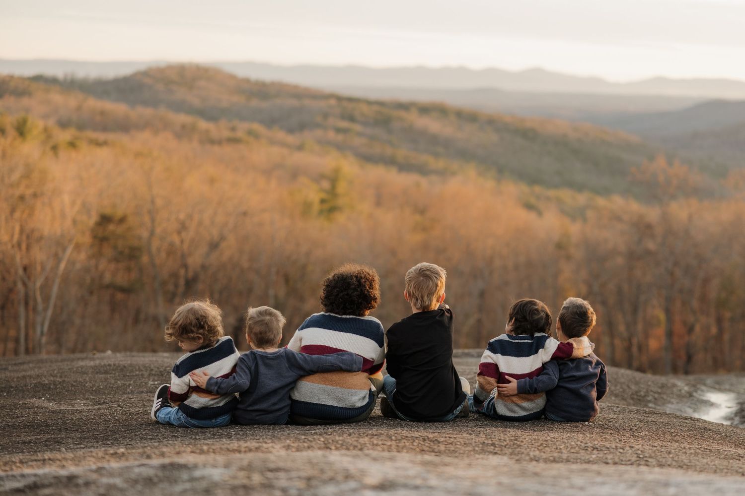 Six children sit together on a rocky overlook, arms around each other, gazing at autumn mountain views.