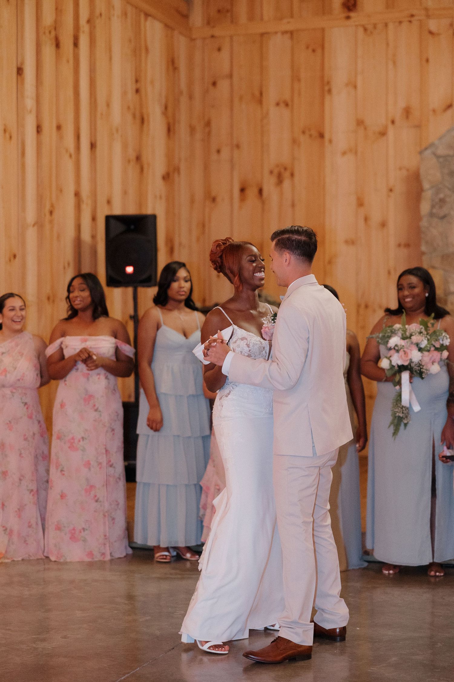 First dance moment in rustic wooden barn with bridal party looking on.