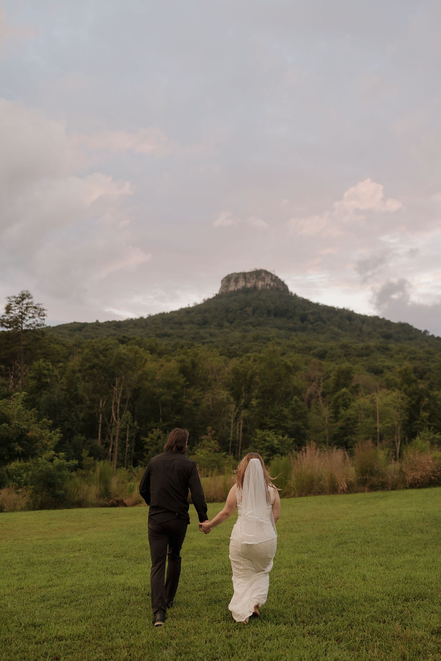 A couple walks away from camera across a grassy field towards a prominent mountain peak.