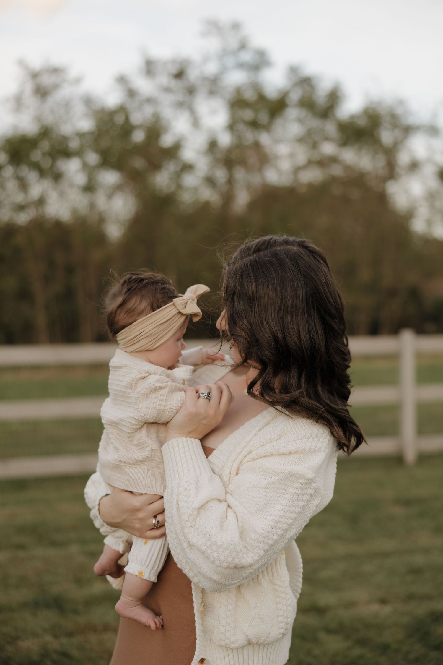 Parent and baby in cream sweaters share tender moments outdoors in a grassy field during golden hour.