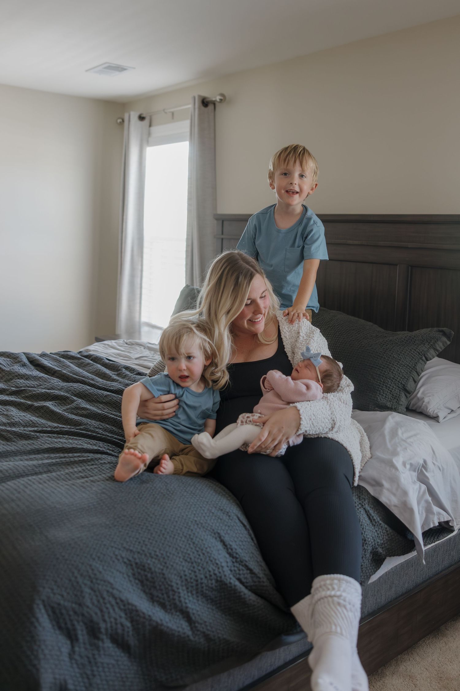 A family sits together on a gray bed with white linens in a bright bedroom with natural lighting.
