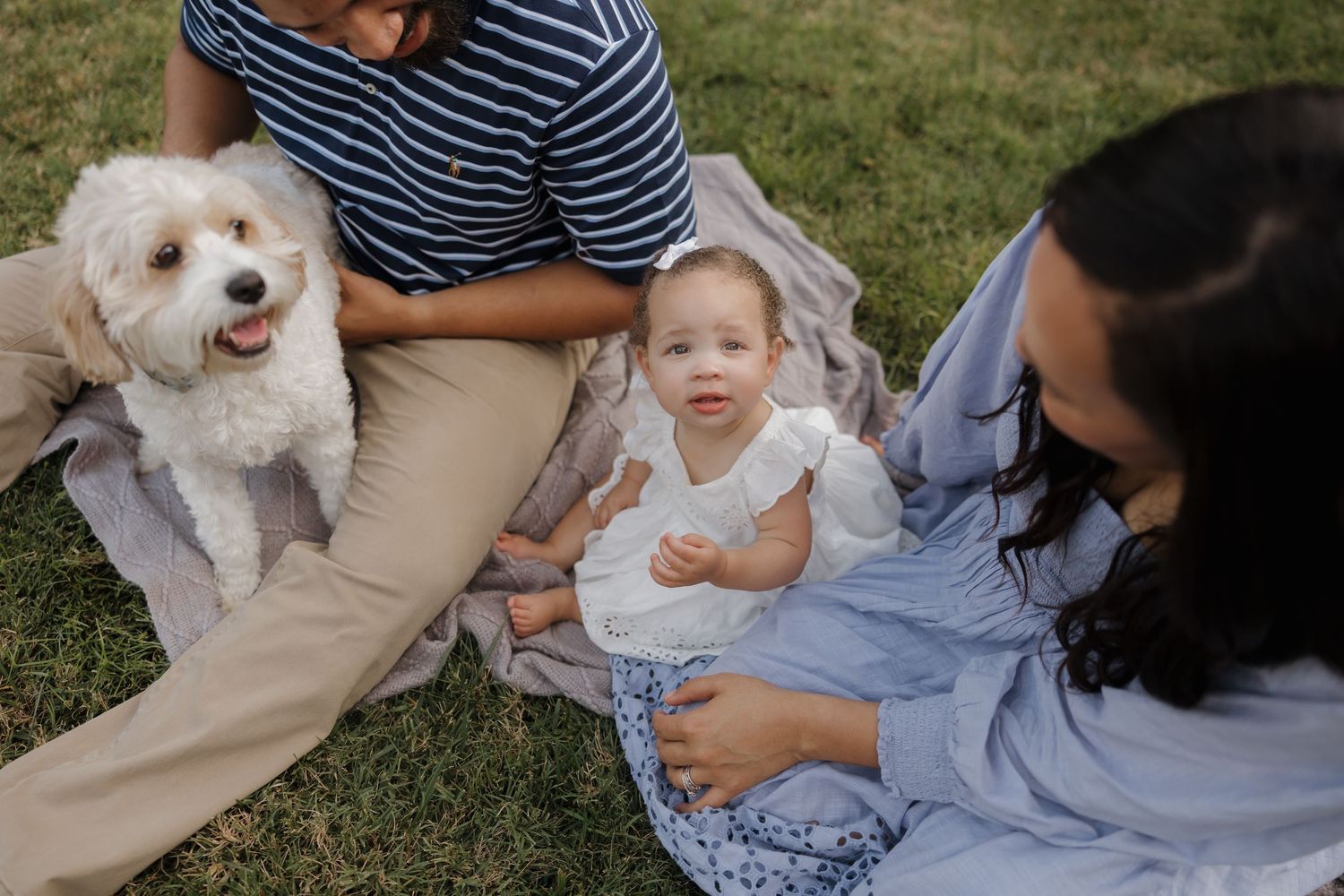 Small white dog and baby in blue dress sit together on grass during outdoor family photoshoot session.