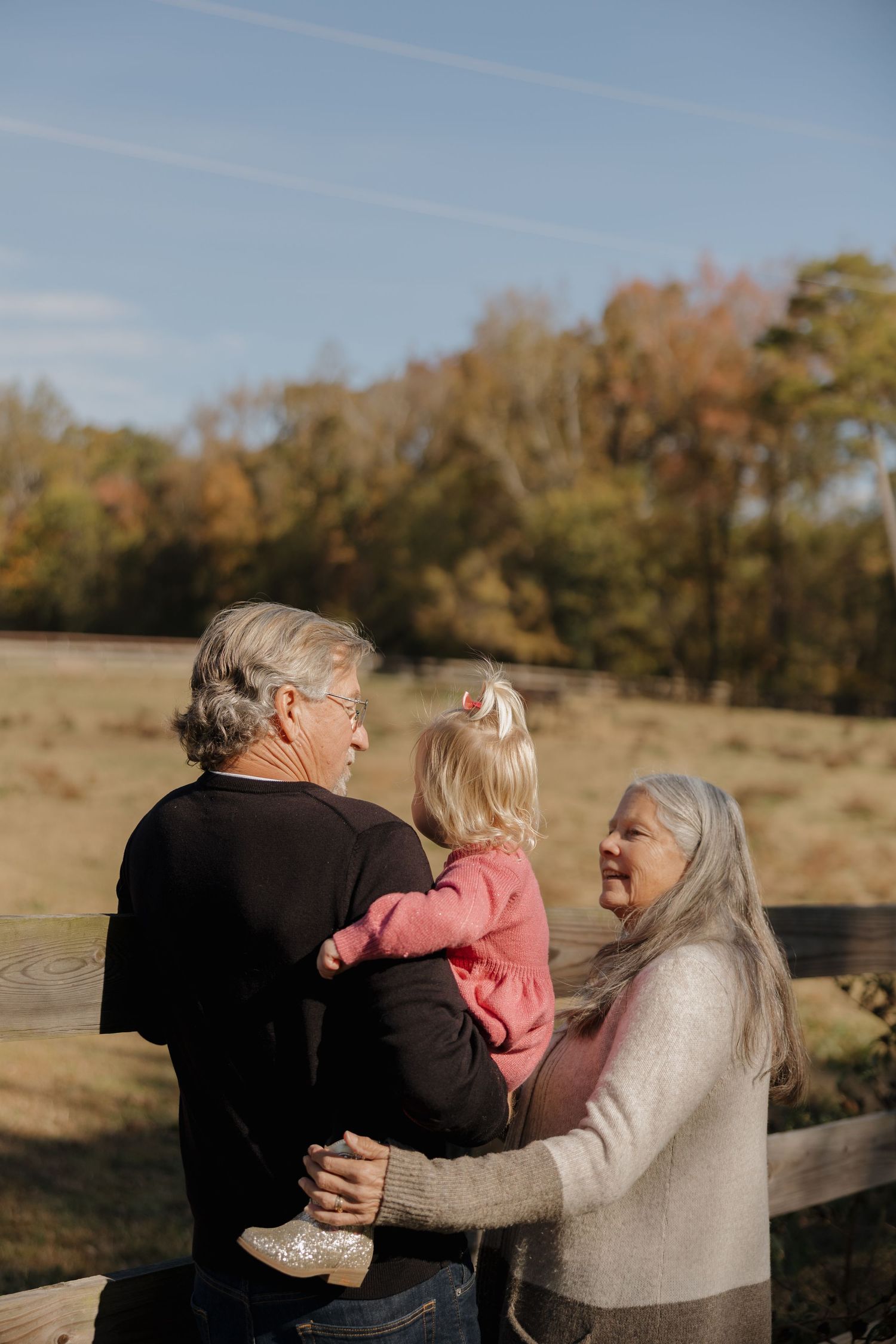 Three people embrace outdoors in a field during golden hour with autumn trees in the background.