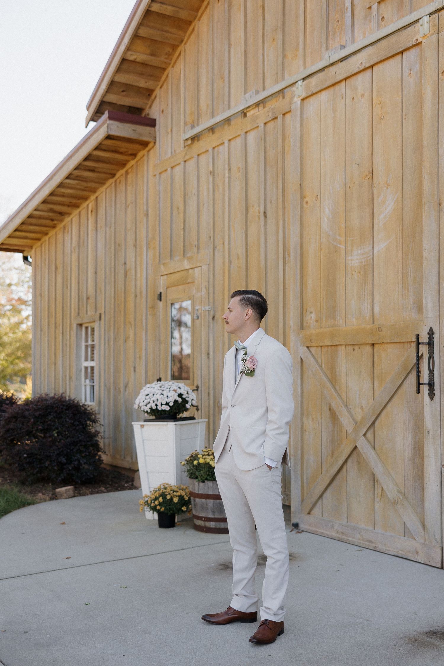 Person in light colored suit stands outside rustic wooden barn.