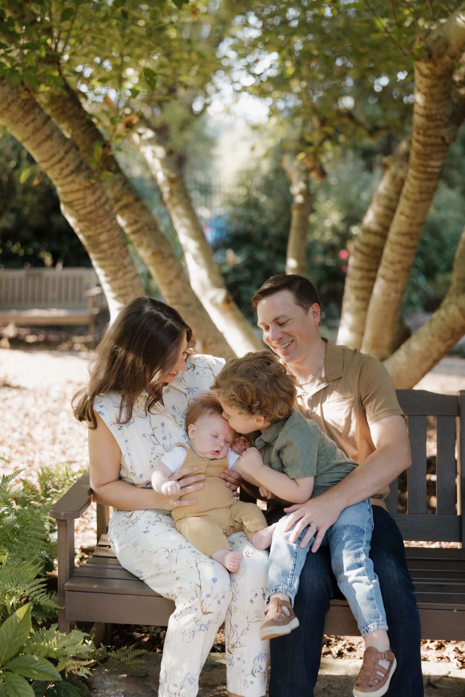 A tender family moment captured on a wooden bench under trees in a park setting with soft natural lighting.