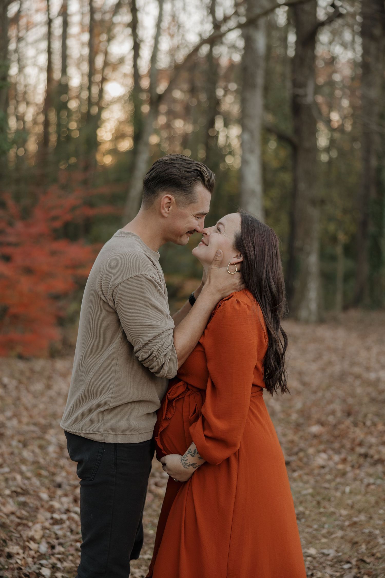 A couple shares an intimate moment in an autumn forest with orange leaves and soft sunlight filtering through trees.
