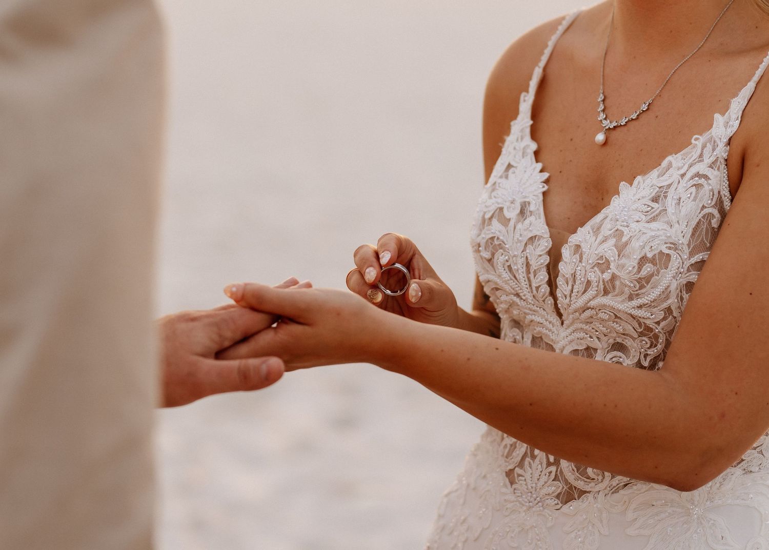 A bride in a lace wedding gown places a ring on her partner's hand during a beach elopement ceremony.
