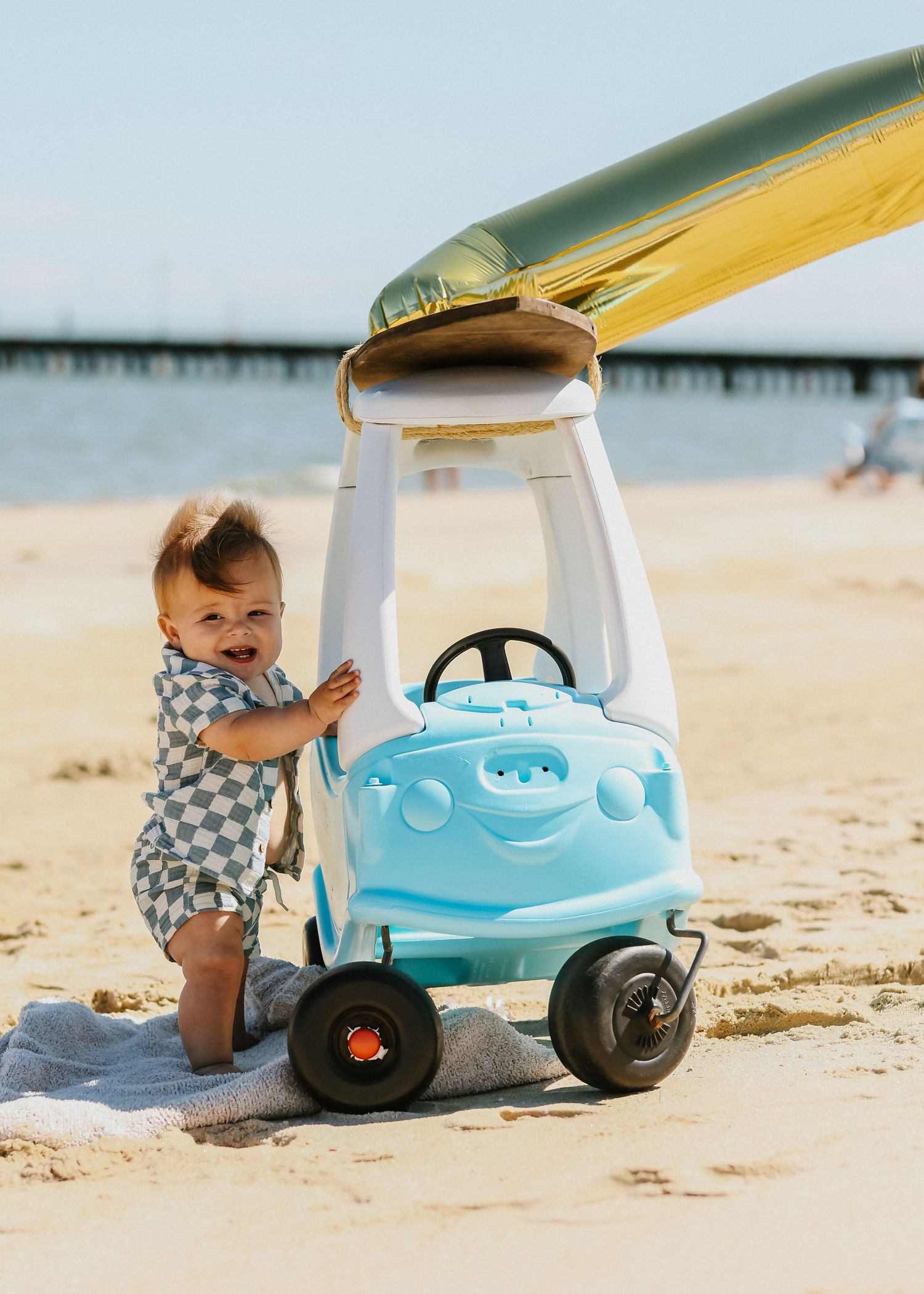 An adorable baby smiles while standing next to a light blue toy car on a sunny sandy beach.