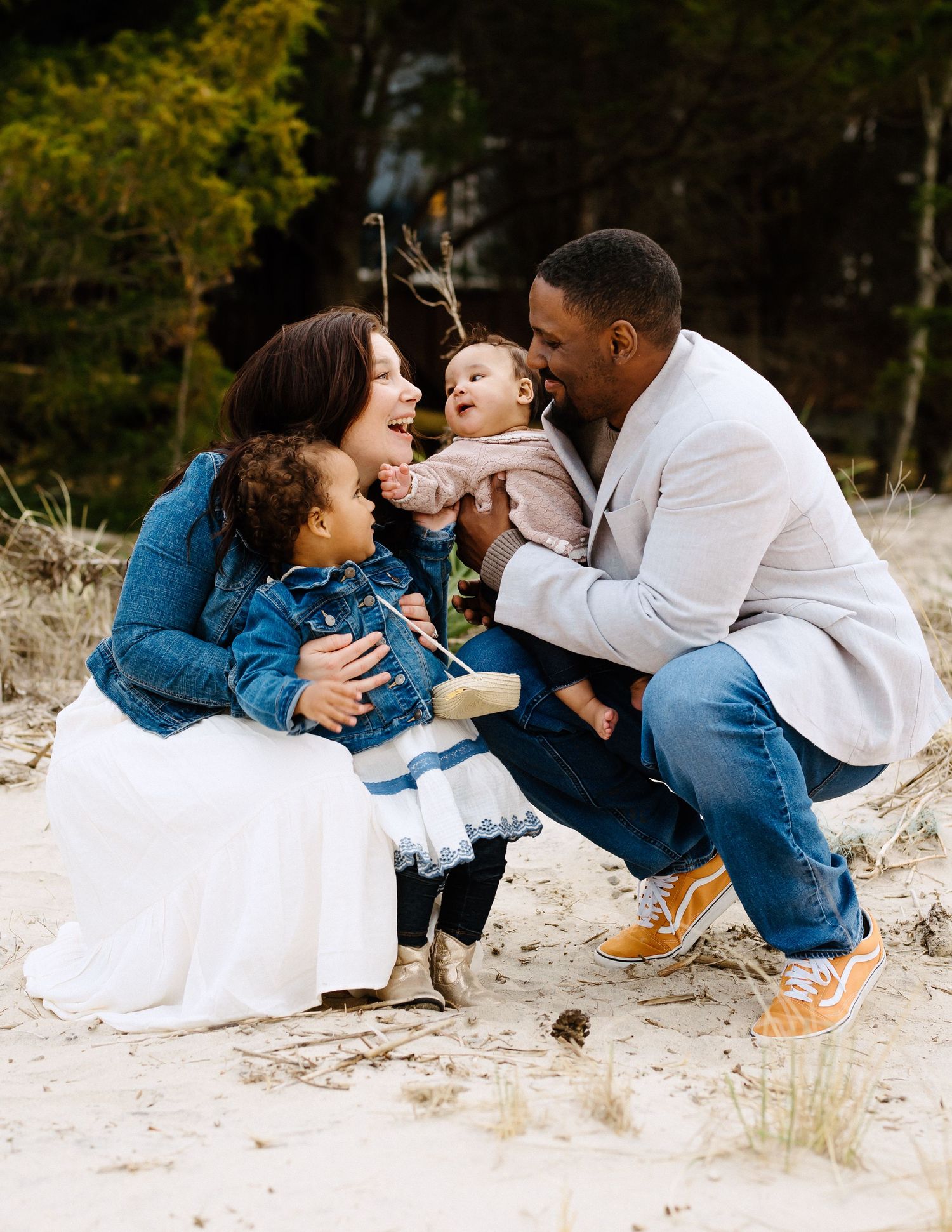 A joyful interracial family with two toddlers laughs together on a sandy beach during a family photo session.