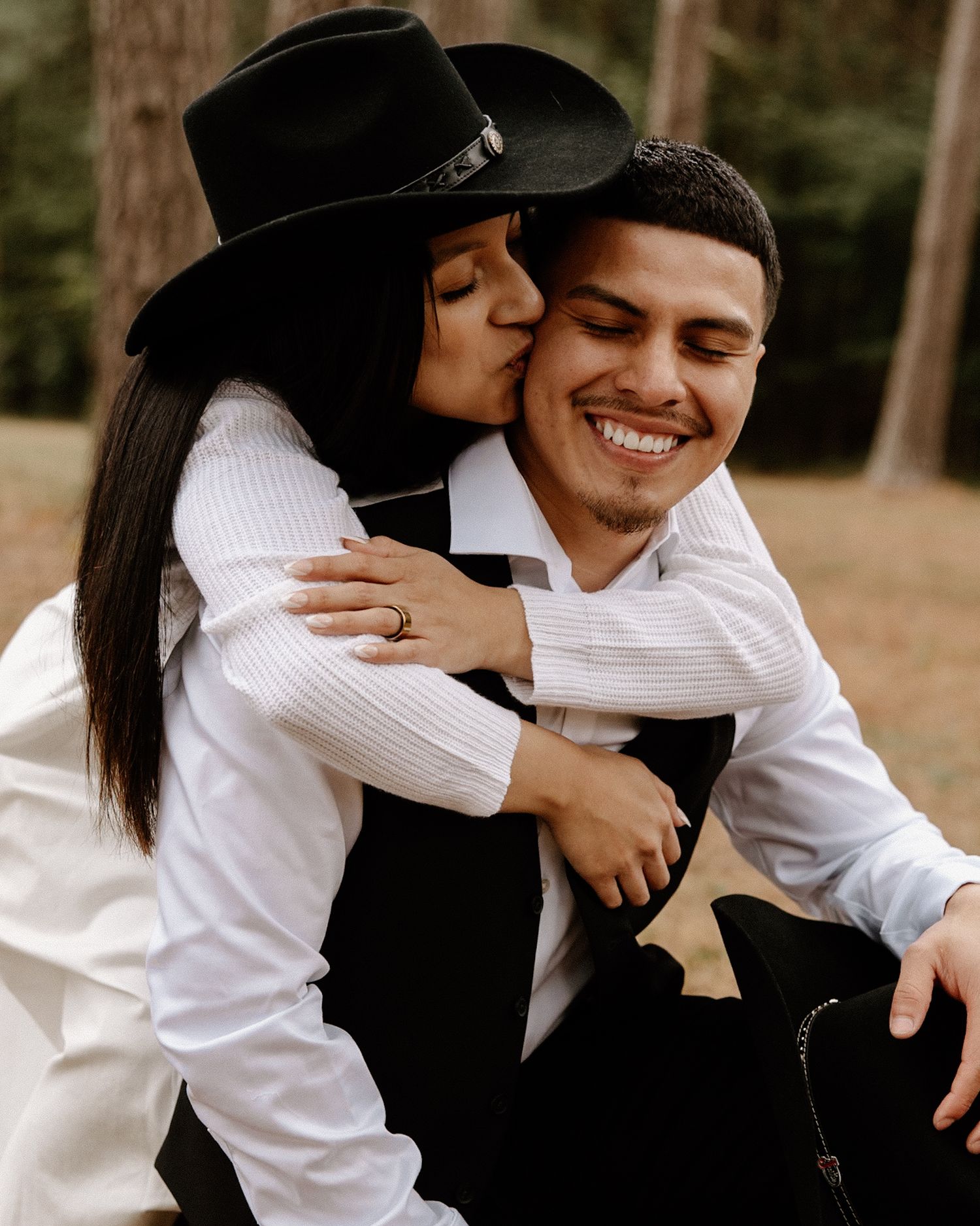 A woman in a black hat kisses her smiling partner on the cheek during a romantic outdoor engagement session.
