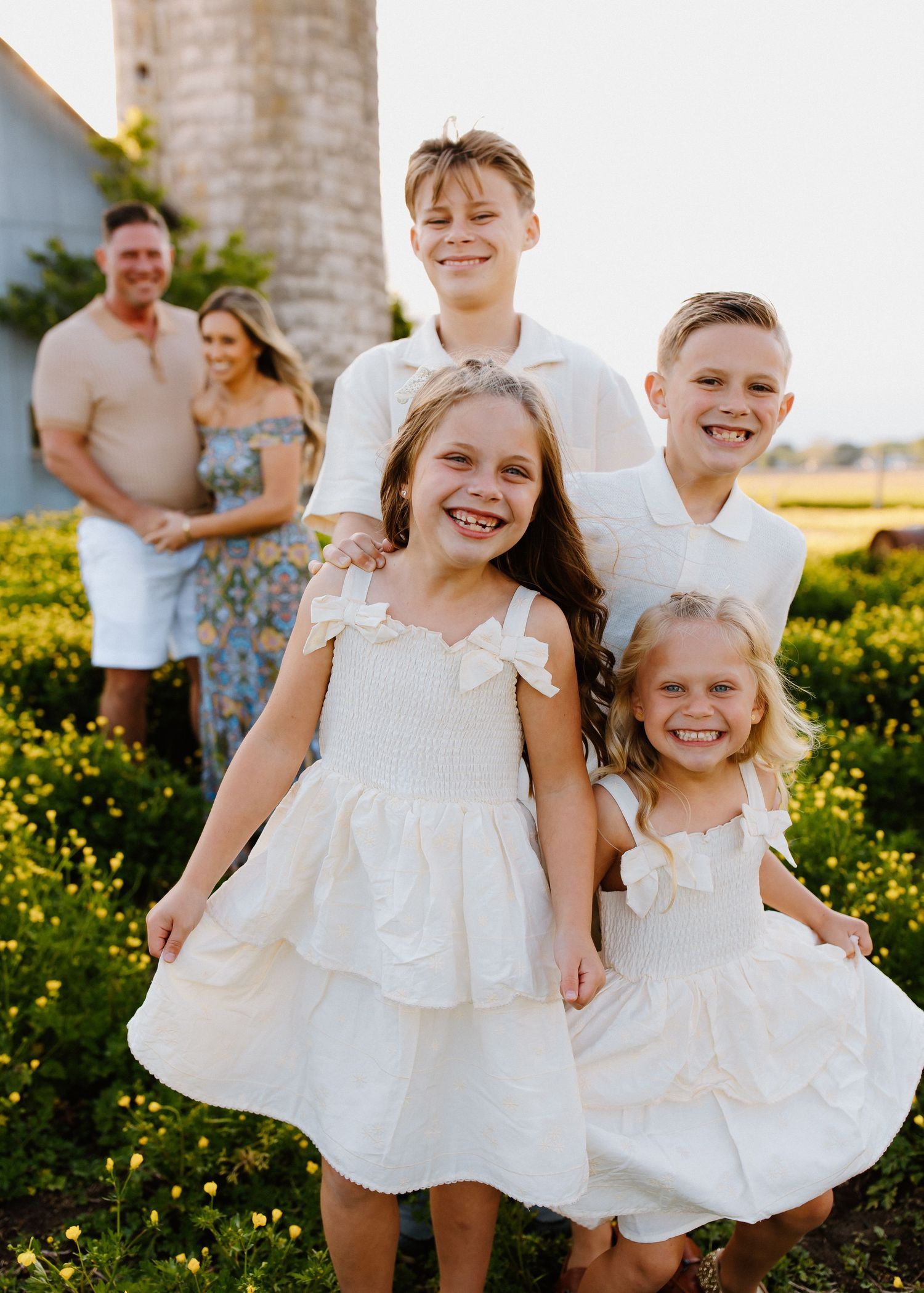 Four happy siblings pose together in white outfits in a sunny flower field during a family portrait session.