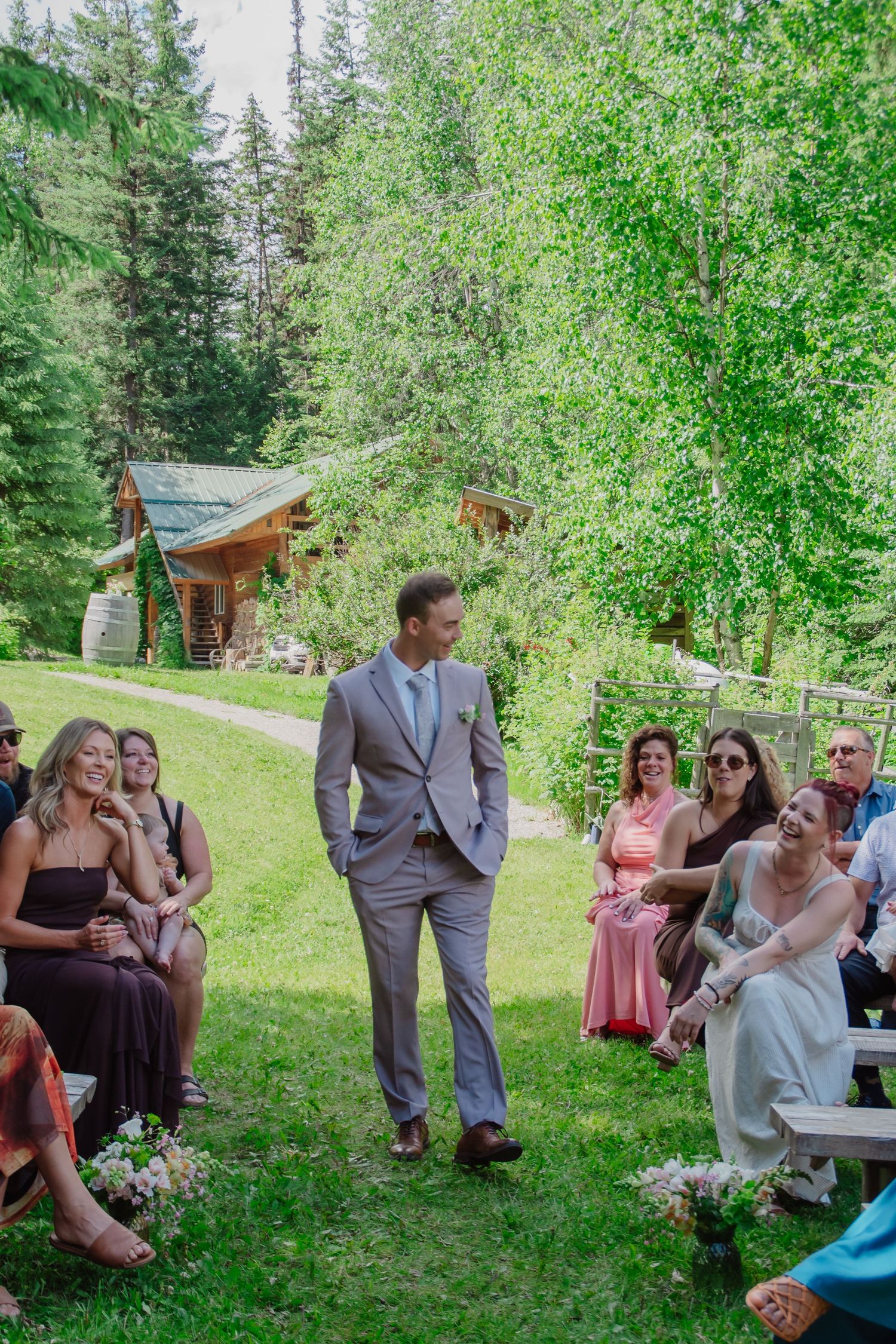 Groom in gray suit walks down outdoor wedding aisle surrounded by seated guests with lush green trees in background.