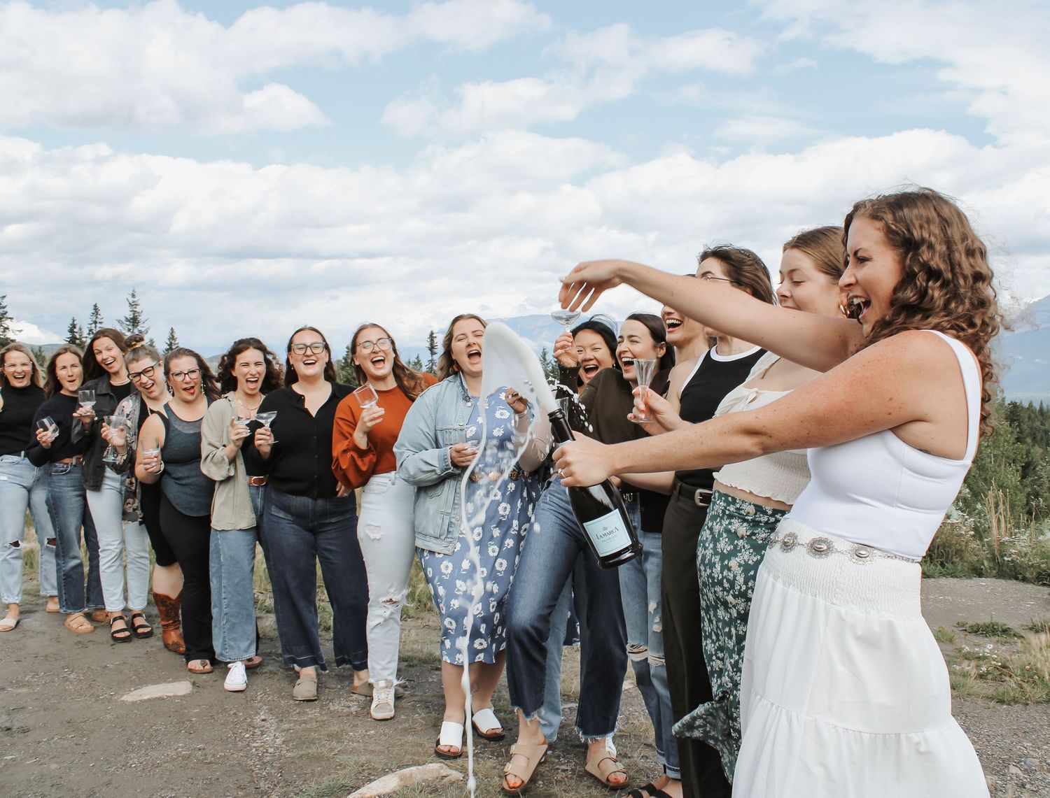 Bride in white dress pops champagne bottle with bridesmaids celebrating outdoors under cloudy sky.