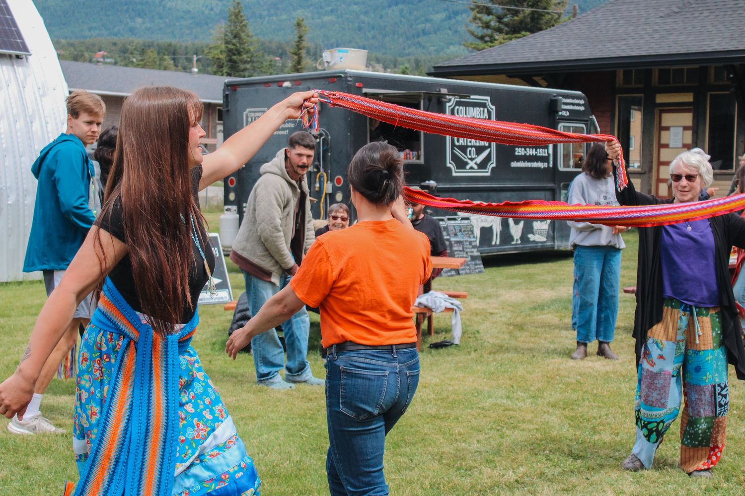 Children and adults playing with ribbons outdoors at a community event near a black food truck on a sunny day.