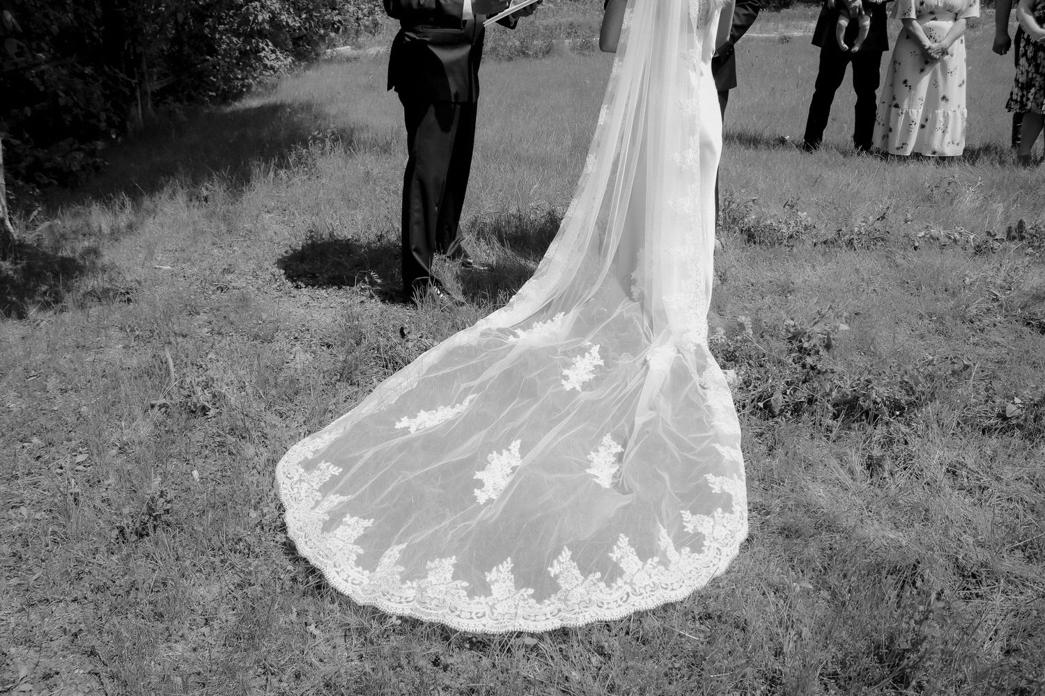 Black and white photo of a bride's lace cathedral wedding veil trailing on grass during outdoor ceremony.