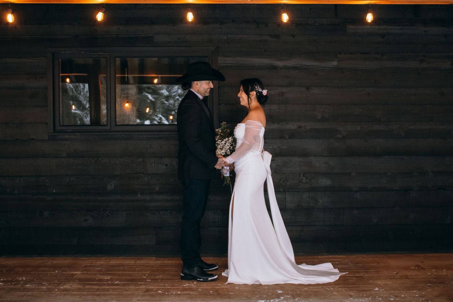 A romantic winter wedding scene with a couple in formal attire standing against a rustic wooden wall with string lights.