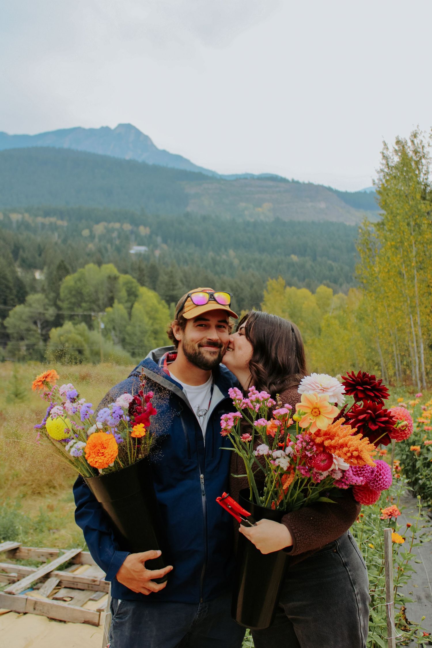 Couple holding colorful flower bouquets with mountain forest backdrop on a cloudy day.